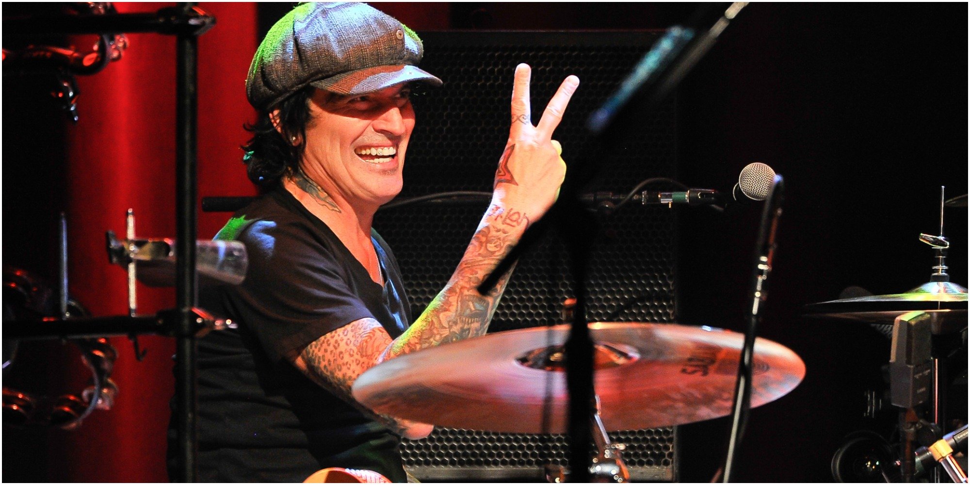 Tommy Lee smiling and holding up two fingers while seated behind the drums.