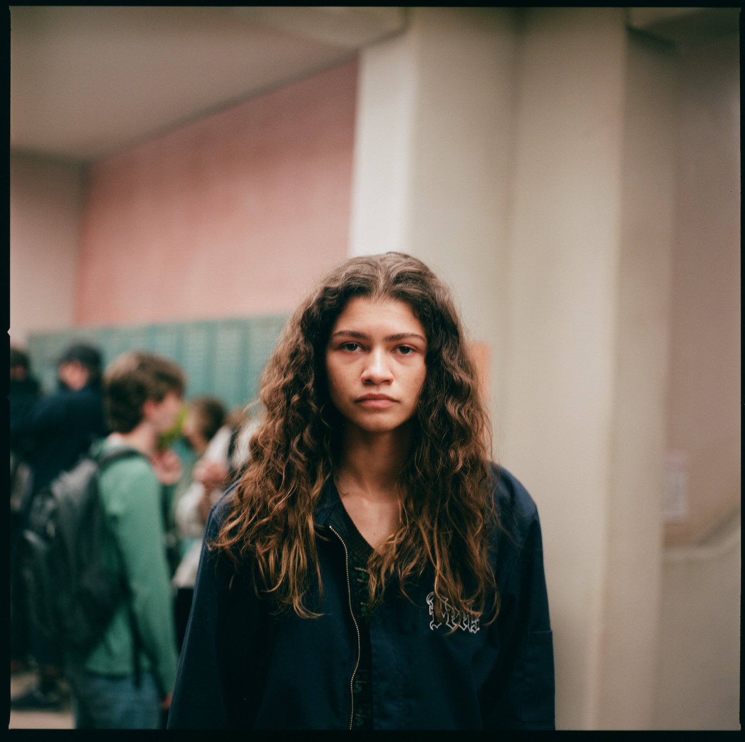 'Euphoria' Season 2 star Zendaya standing in front of lockers at a school wearing a blue zip-up hoodie in a production still.