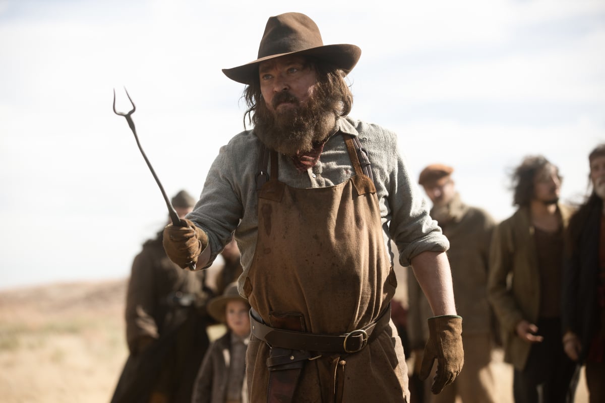 James Jordan as Cookie of the Paramount+ original series 1883. Cookie wears a brown apron and cowboy hat and brandishes a long skewer.