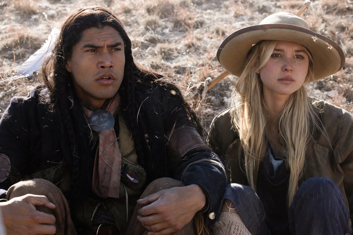 Martin Sensmeier as Sam and Isabel May as Elsa of the Paramount+ original series 1883. Sam and Elsa sit beside each other on the ground.  