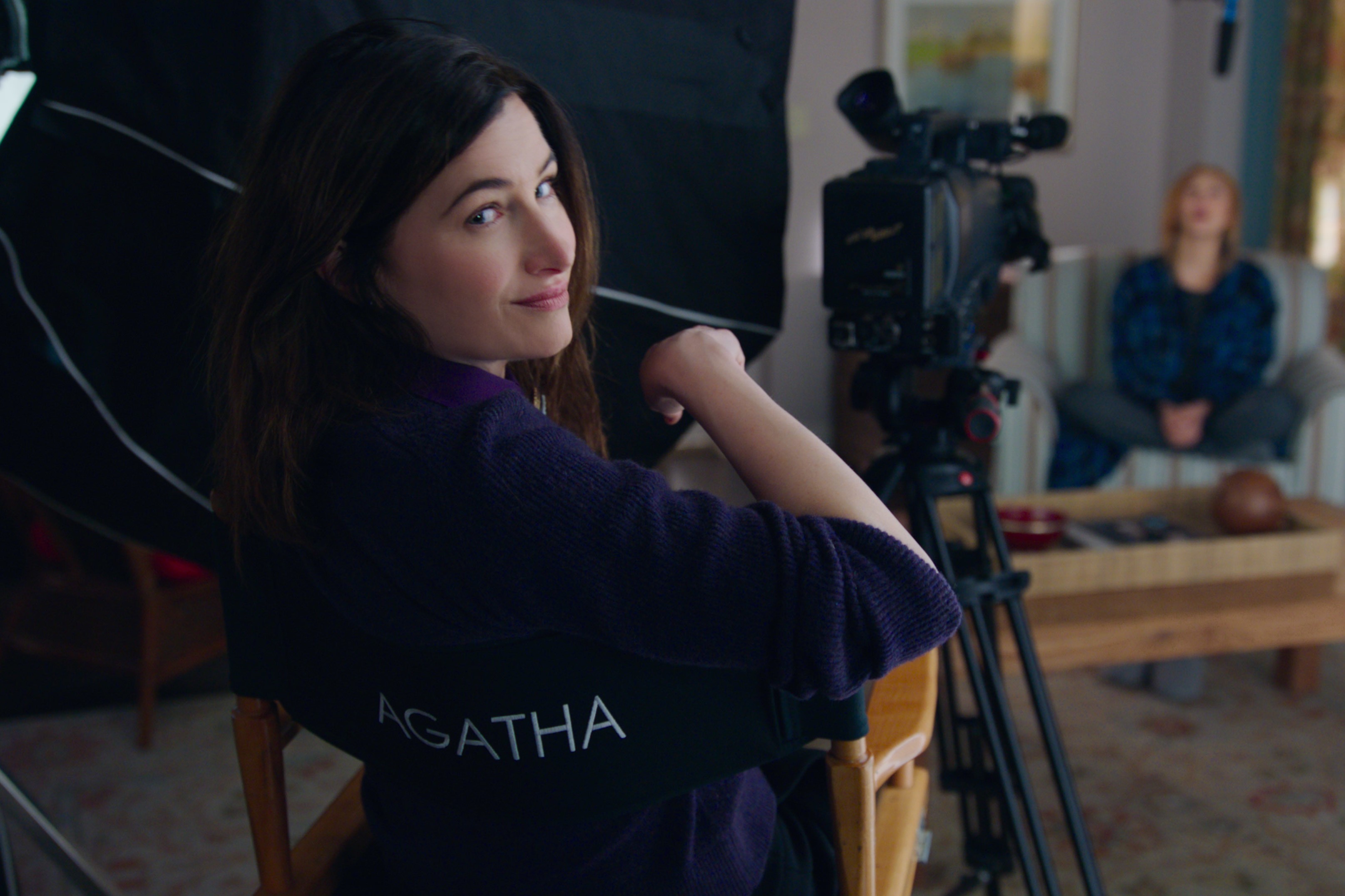 Kathryn Hahn, in character as Agatha Harkness, who fans want to see in 'Doctor Strange 2' after seeing the trailer, wears a purple sweater and sits in a director's chair that reads "Agatha" on the back. Agatha sits behind a camera that records Wanda in the background.