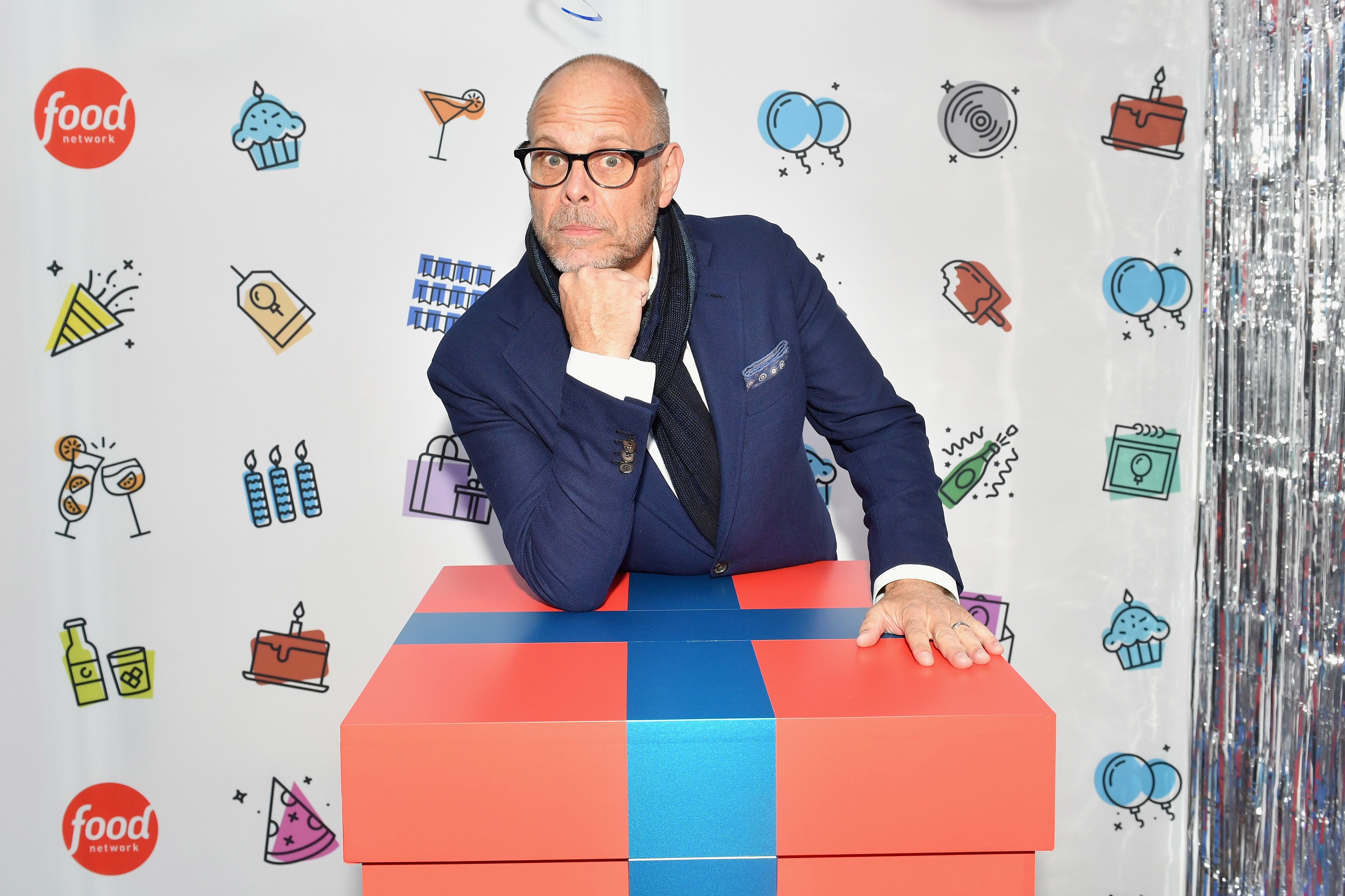 Chef Alton Brown poses with elbow on a giant wrapped gift at a 2018 Food Network event.