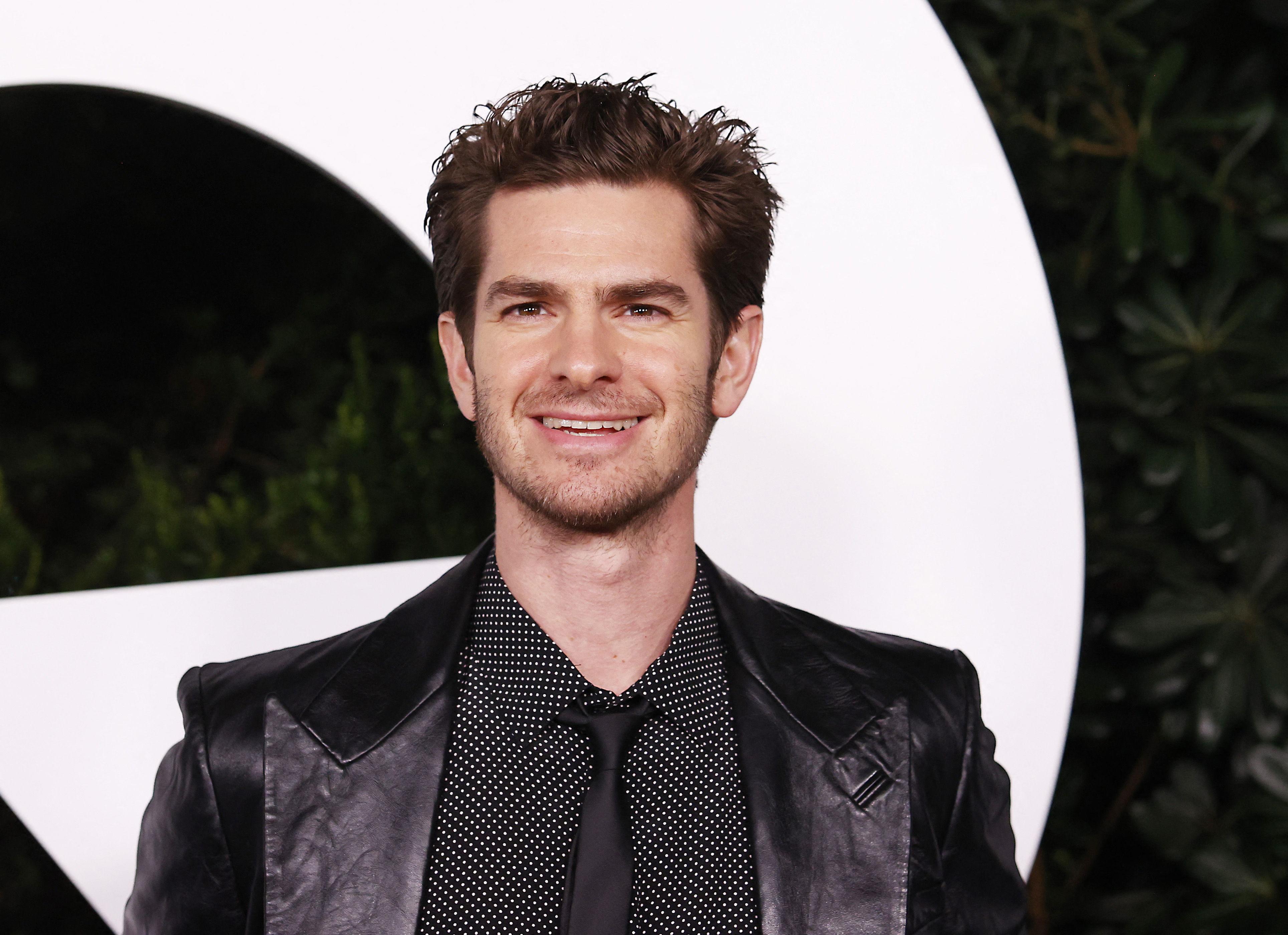 Andrew Garfield, who starred alongside Tobey Maguire in 'Spider-Man: No Way Home,' wears a black leather suit jacket over a black button-up shirt with small white polka dots and a black tie.