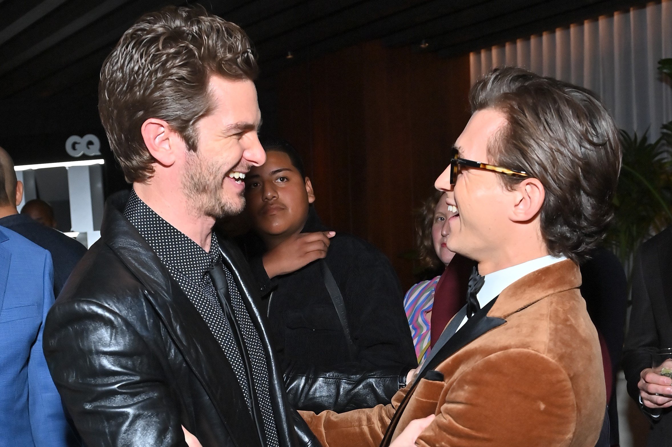 'Spider-Man: No Way Home' stars Andrew Garfield and Tom Holland smile at one another as they embrace. Garfield wears a black leather suit jacket over a black button-up shirt with white polka dots and a black tie. Holland wears a brown velvet suit jacket over a white button-up shirt and black tie.