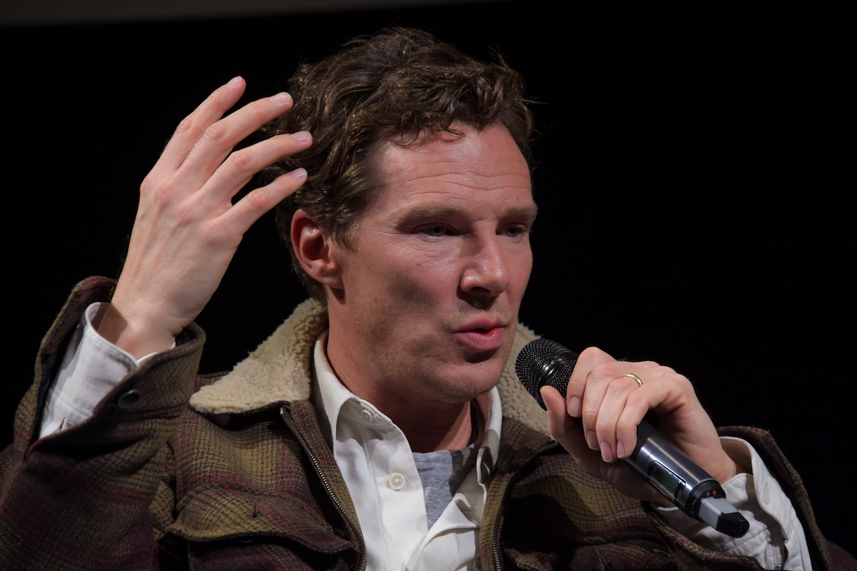 Benedict Cumberbatch holds a microphone to his mouth while he raises his hand