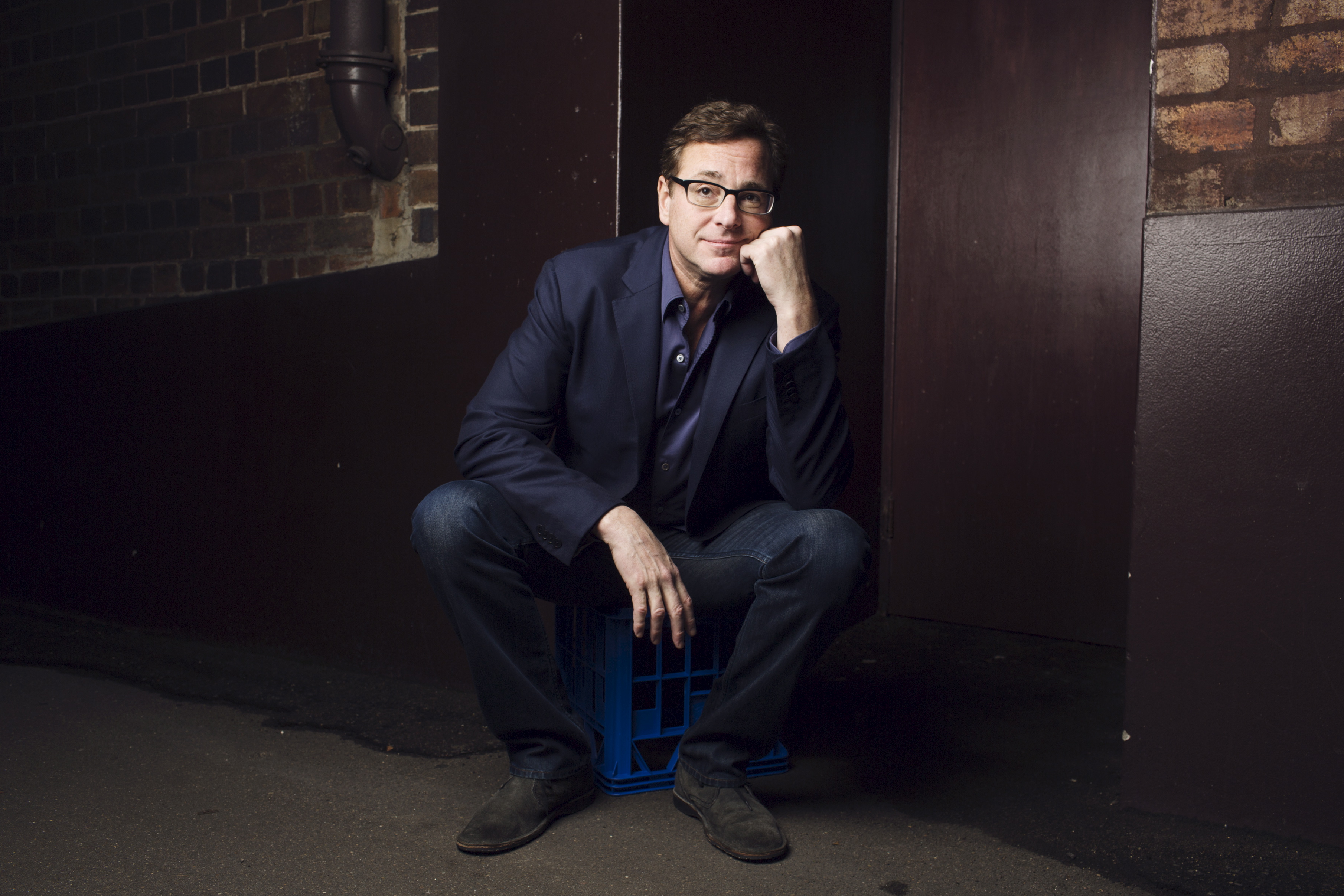Bob Saget sits down with his hand under his chin in front of a dark background. 