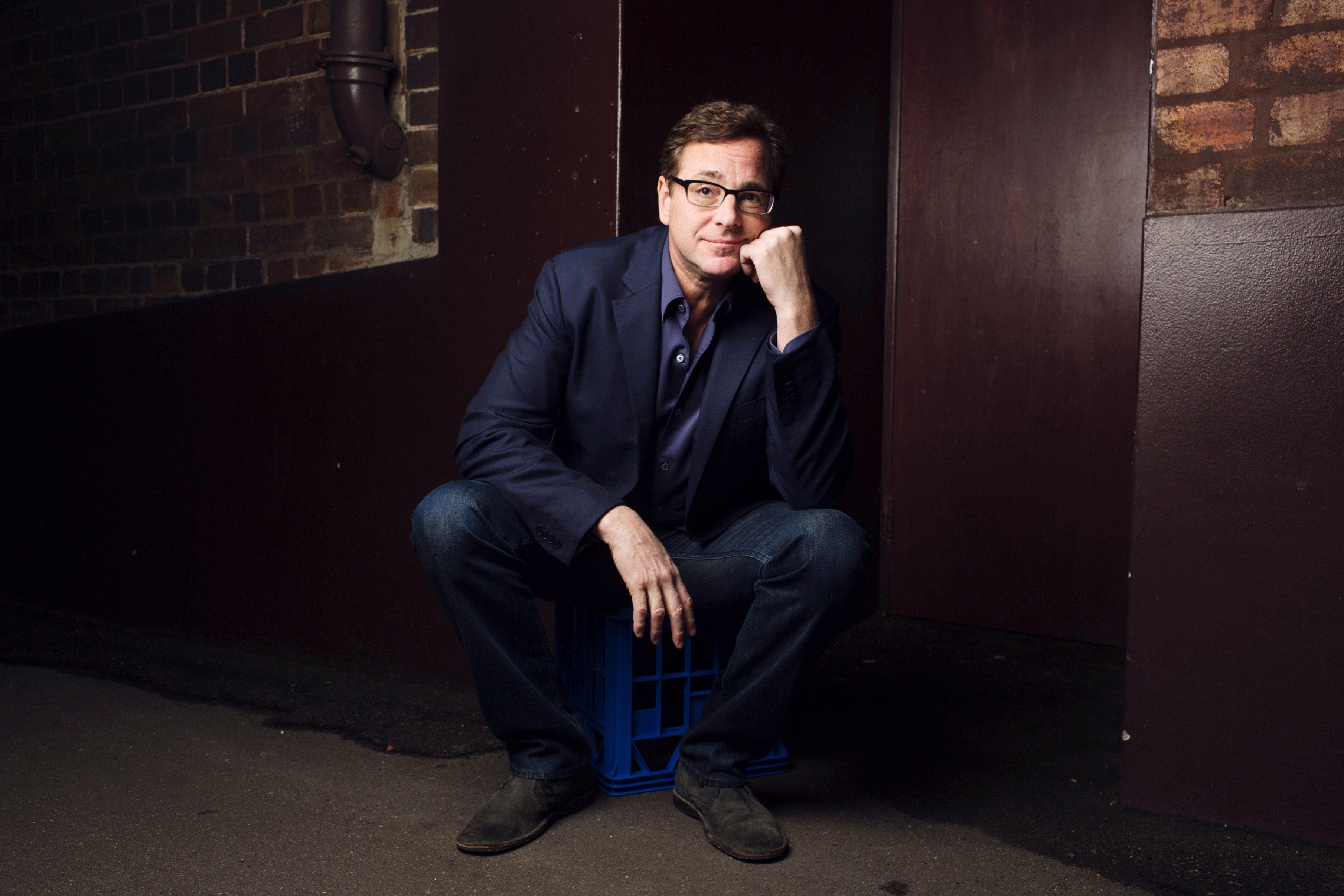 Bob Saget sits down with his hand under his chin in front of a dark background.
