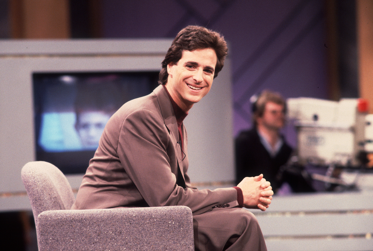 Bob Saget sits on a couch and smiles.