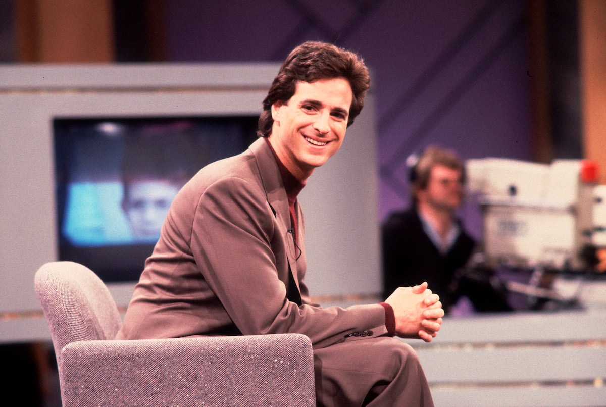 Bob Saget sits on a couch and smiles.