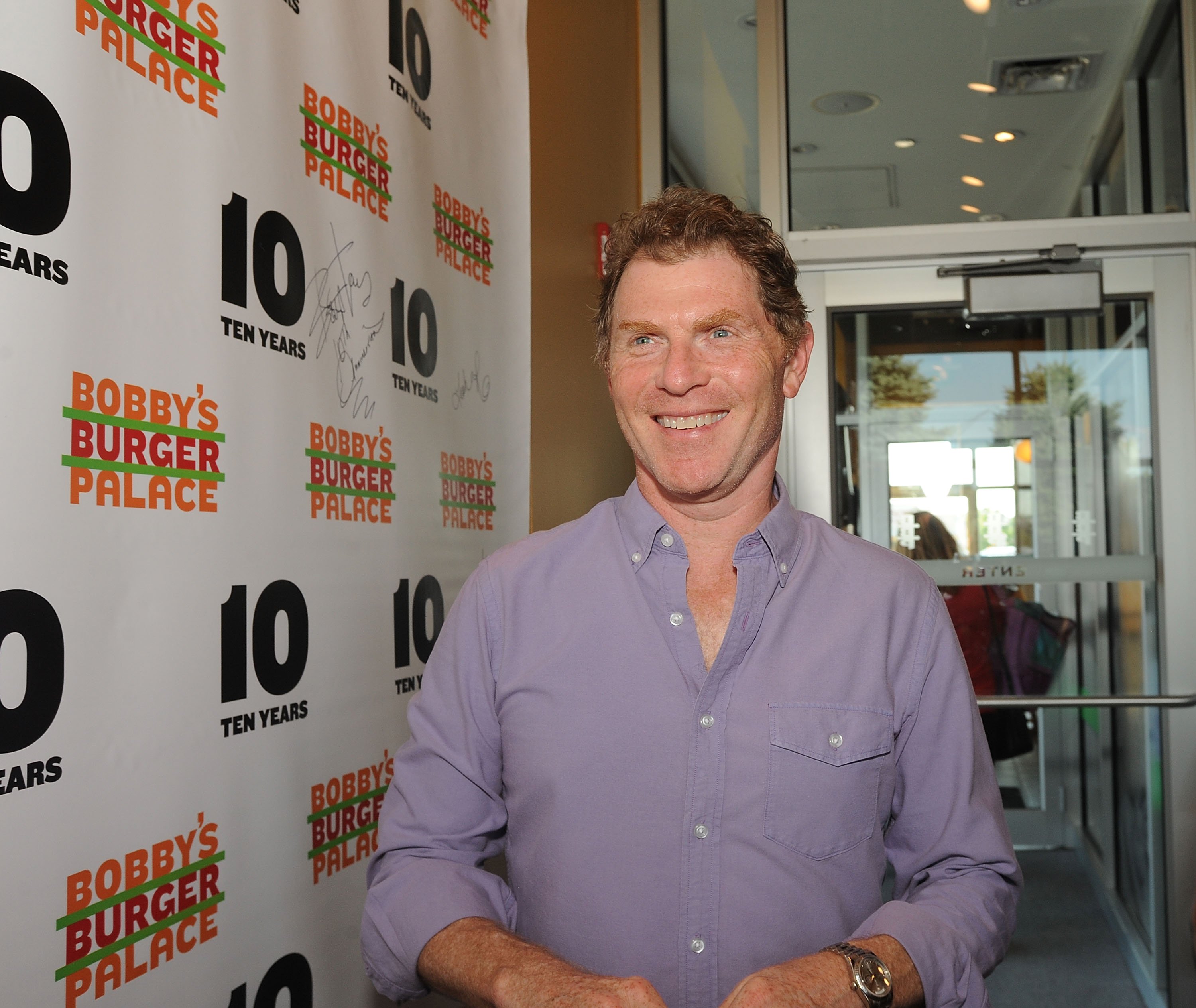 Food Network chef Bobby Flay is wearing a long-sleeve button-down shirt as he visits a Bobby's Burger Palace restaurant in 2018.