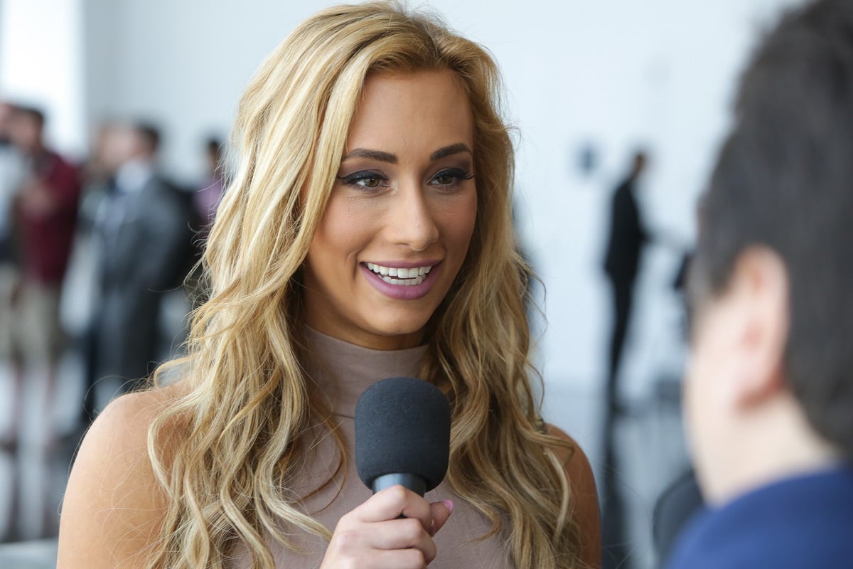 WWE star Carmella wearing a beige dress and holding a microphone.