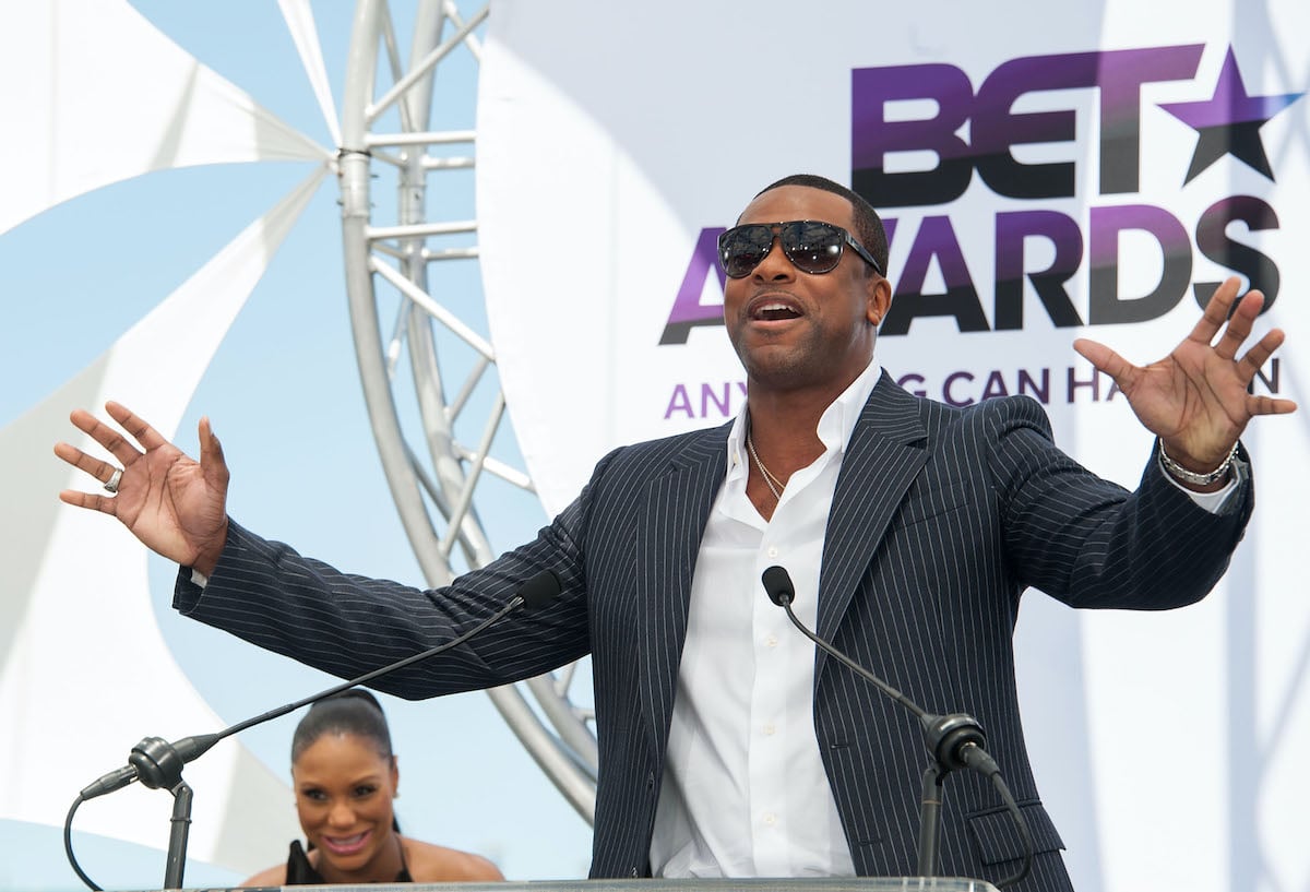 Chris Tucker wears a striped jacket and sunglasses as he speaks at a podium