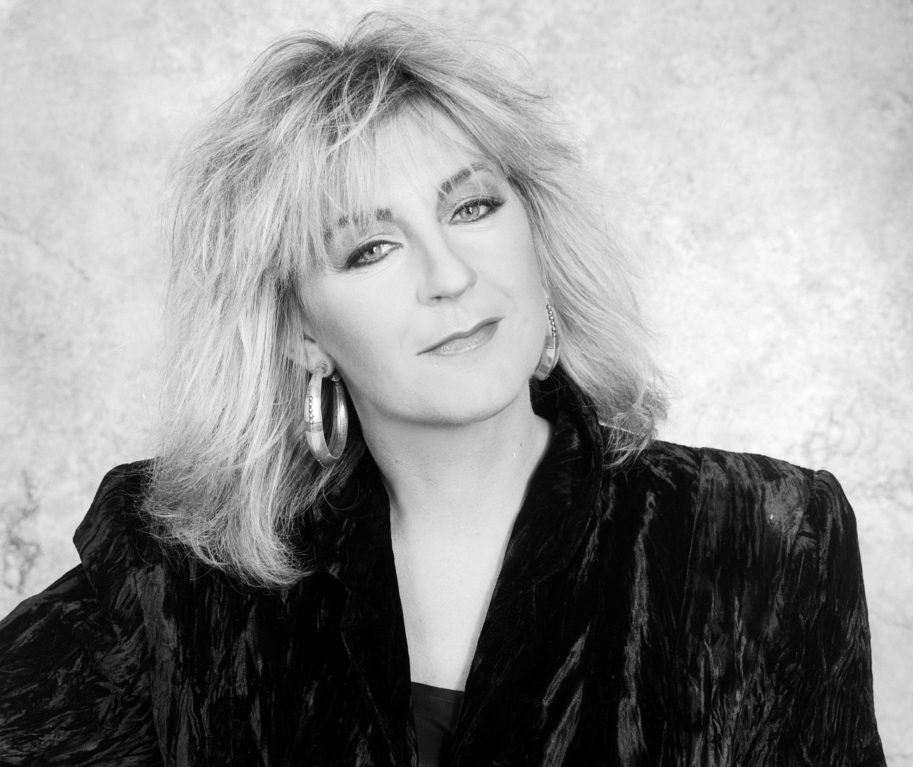 A black and white photo of Christine McVie wearing a black jacket and hoop earrings.