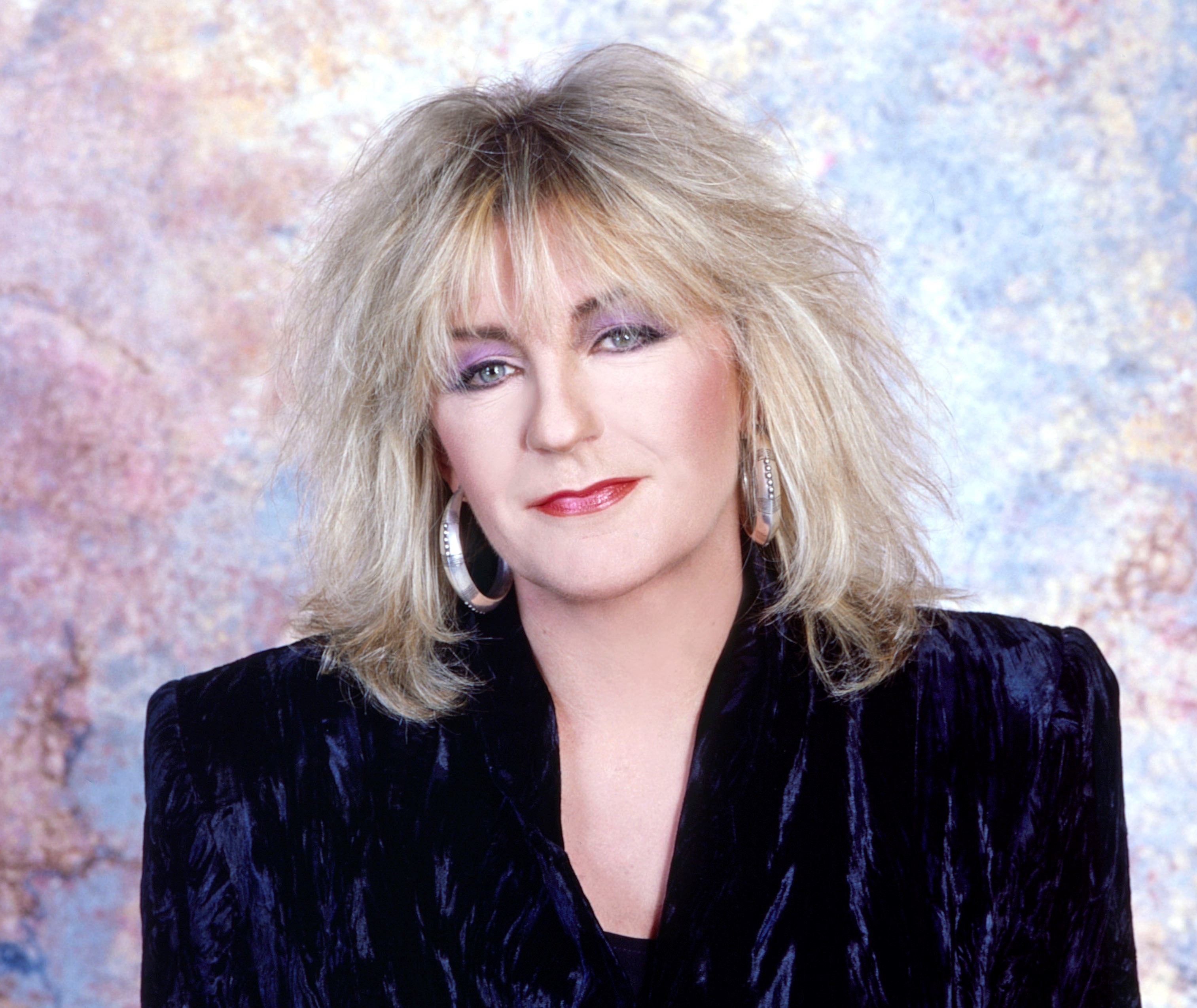 Christine McVie wears a black shirt and silver earrings against a multicolored background.