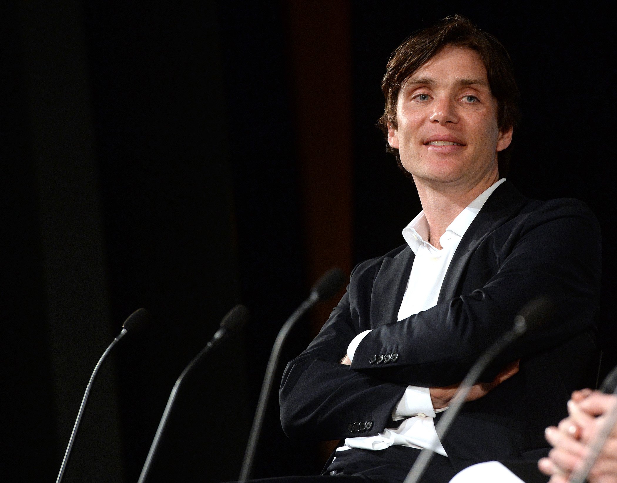 Thomas Shelby actor Cillian Murphy from 'Peaky Blinders' Season 6 sitting with his arms crossed and smiling against a black background