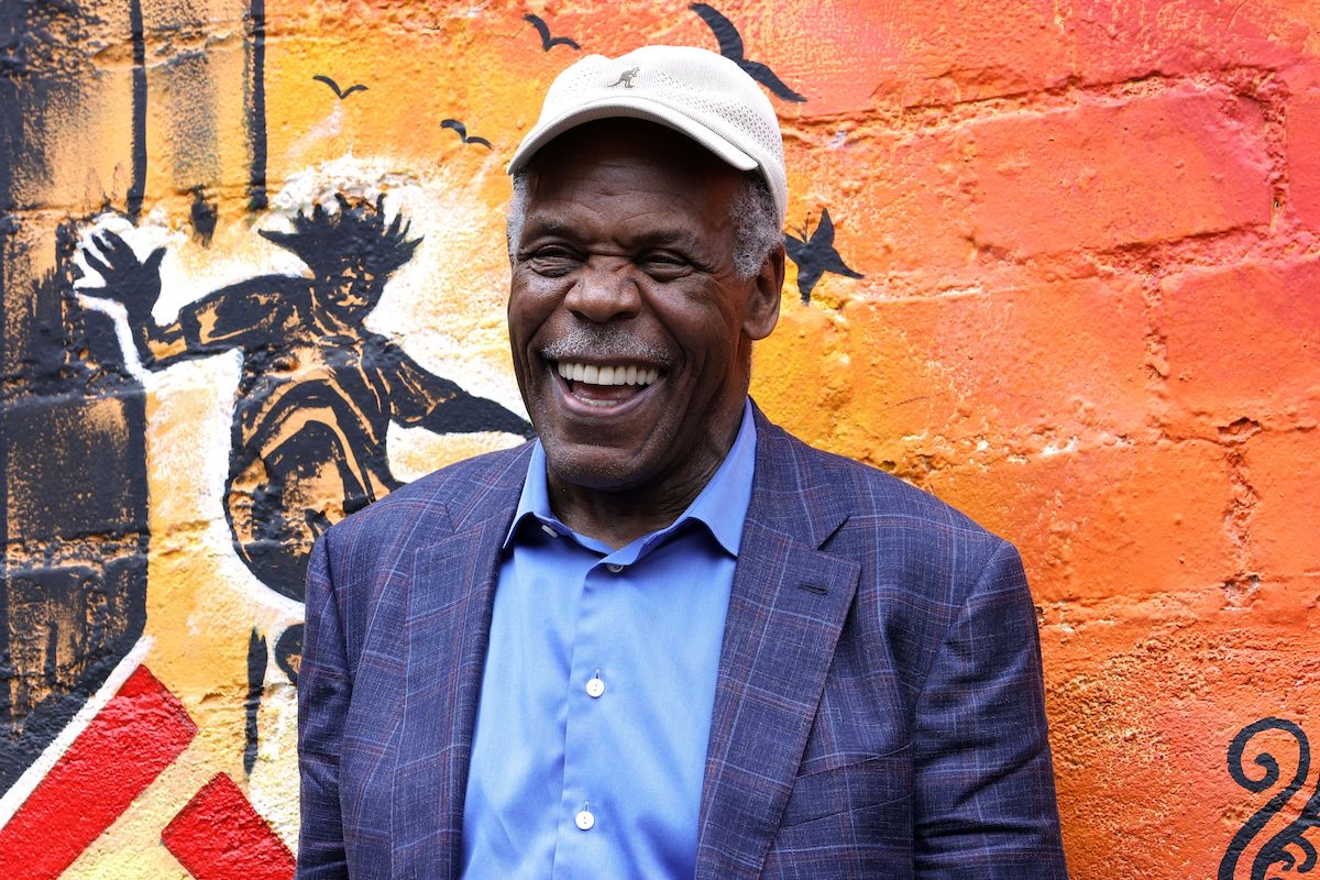 Danny Glover laughs while wearing a blue shirt and jacket in front of an orange wall