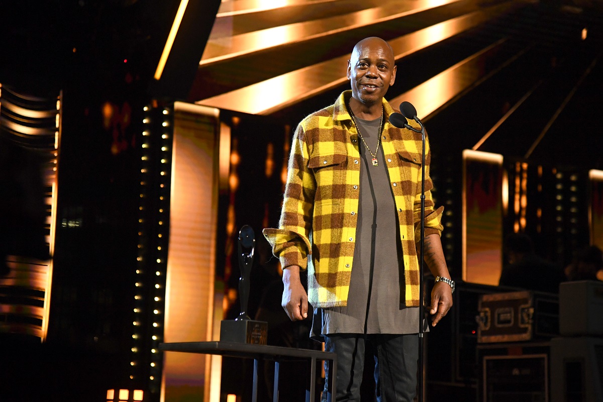 Dave Chappelle smiling while wearing a yellow shirt in front of a microphone on stage.