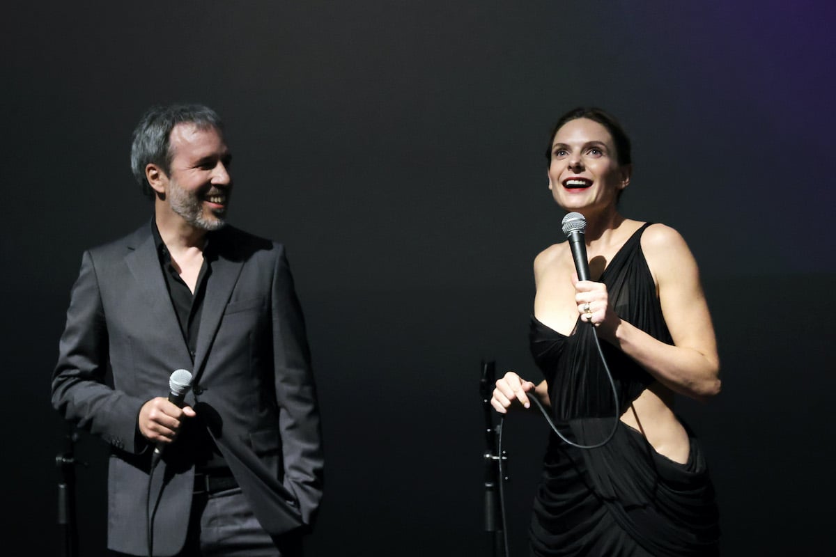 Denis Villeneuve wears a gray jacket and Rebecca Ferguson in a black dress speak onstage