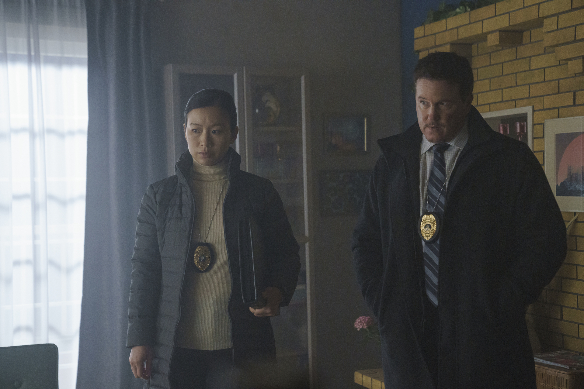 Annie Chang and Lochlyn Munro as Sophie Song and Larry Fitzgibbon in 'Peacemaker.' They're standing next to one another and looking down at something.