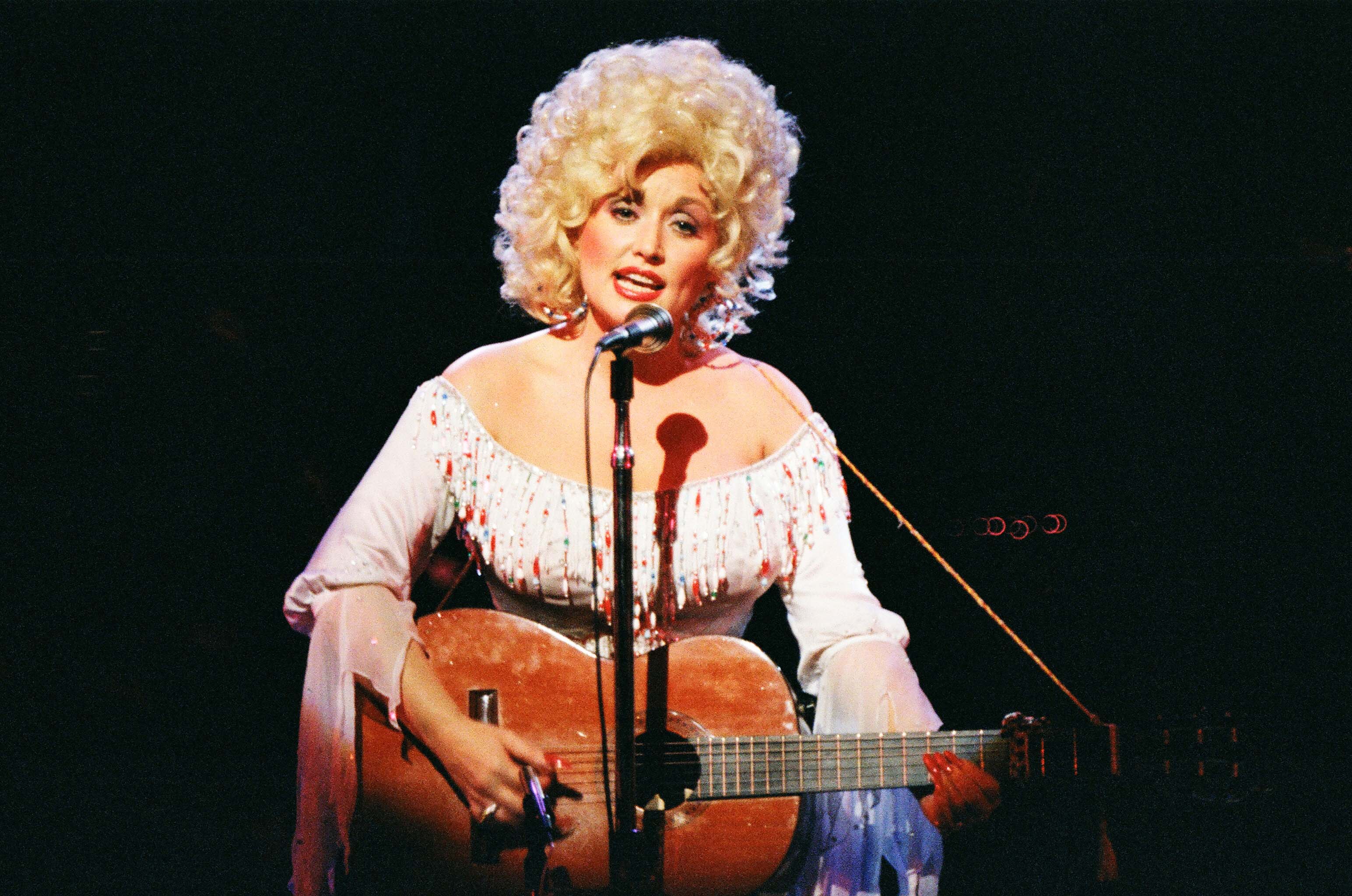 Dolly Parton wears a white rhinestoned dress and sings into a microphone. She holds a guitar.