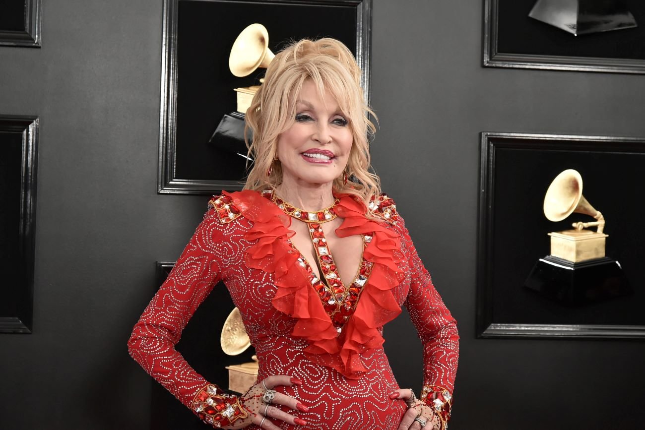 Dolly Parton wears a red dress and stands with her hands on her hips in front of a background with Grammy statues.