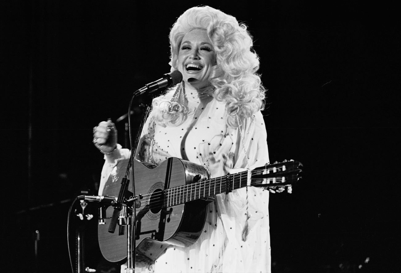 A black and white photo of Dolly Parton holding a guitar and smiling in front of a microphone. 