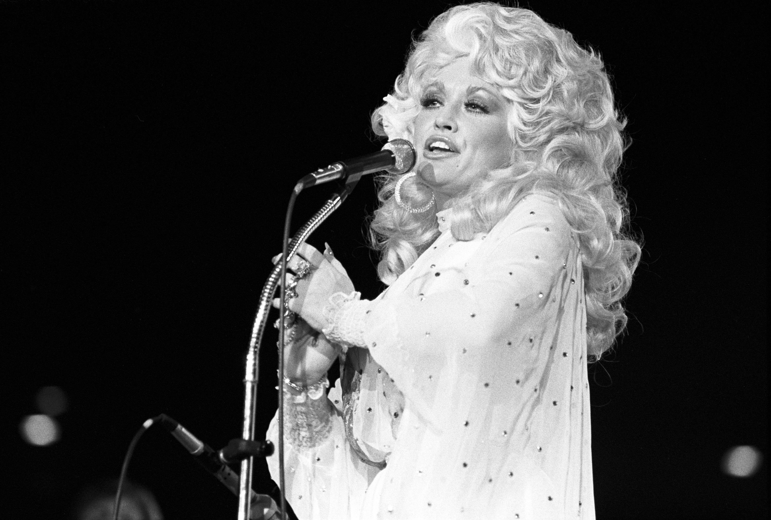 A black and white photo of a '70s Dolly Parton standing in front a microphone.