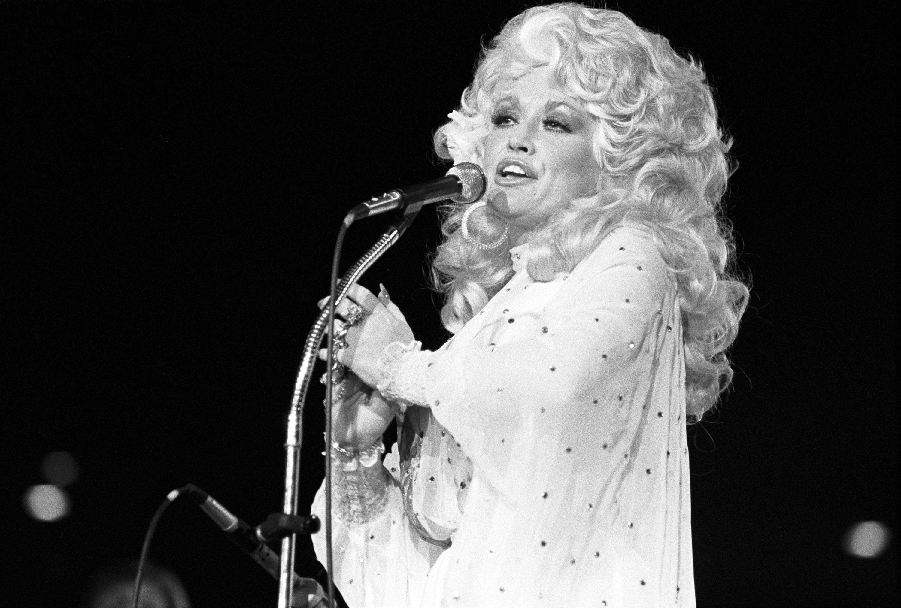 A black and white photo of a '70s Dolly Parton standing in front a microphone.