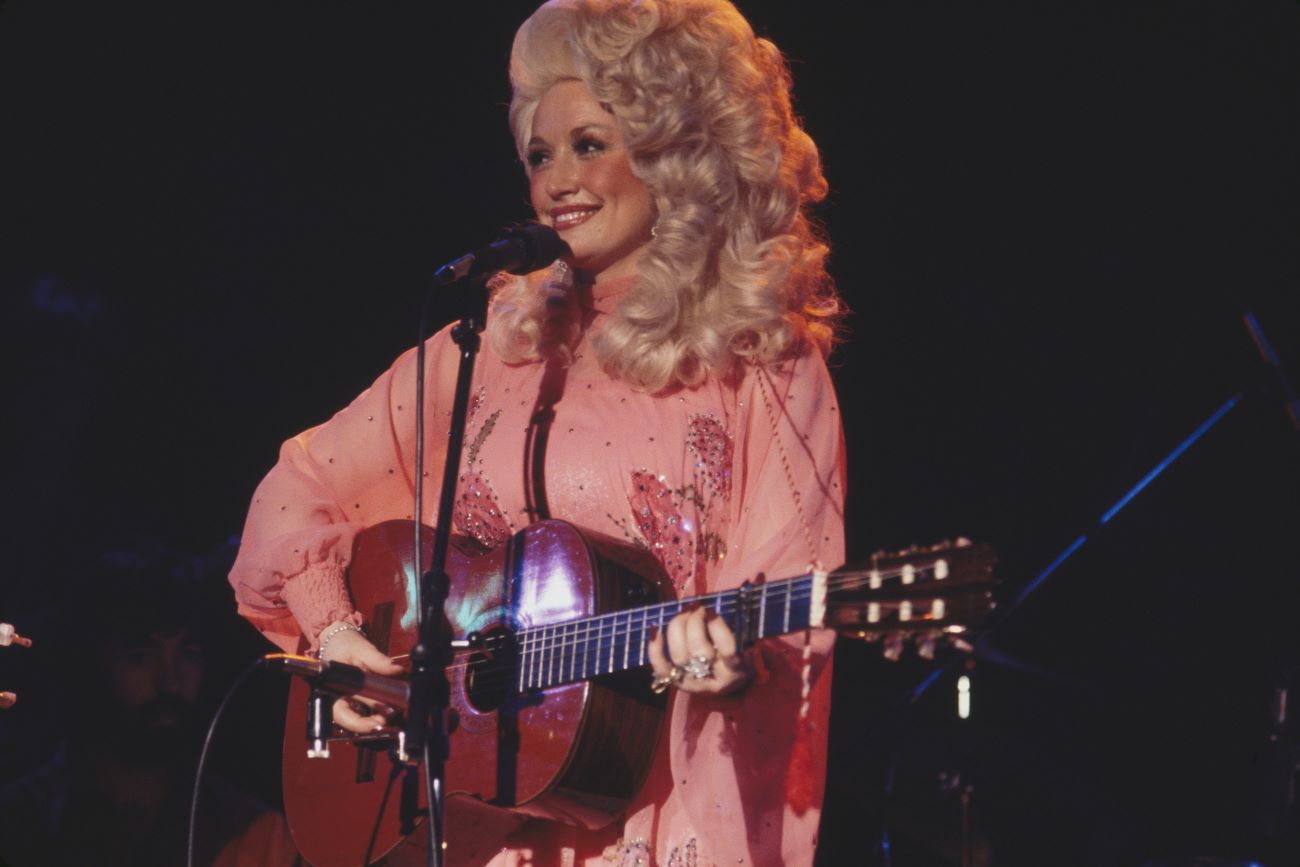 Dolly Parton wears a pink dress and strums a guitar in front of a microphone.