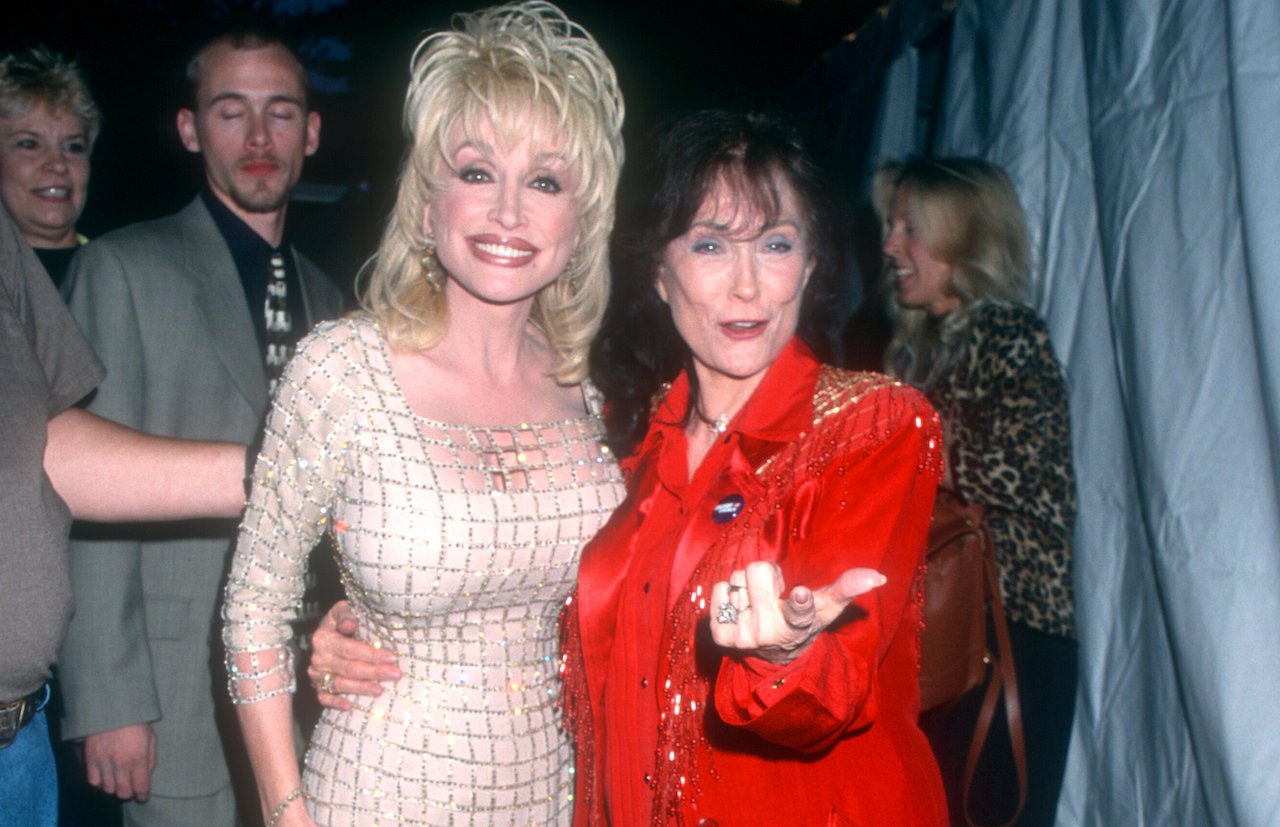 Dolly Parton in a light colored dress standing with Loretta Lynn in red