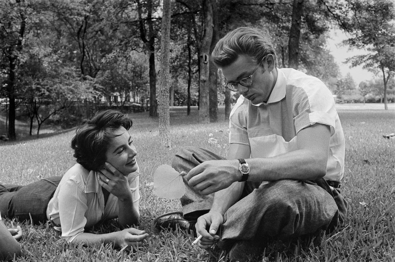 James Dean usa anteojos y se sienta con las piernas cruzadas en el césped.  Elizabeth Taylor descansa junto a él.