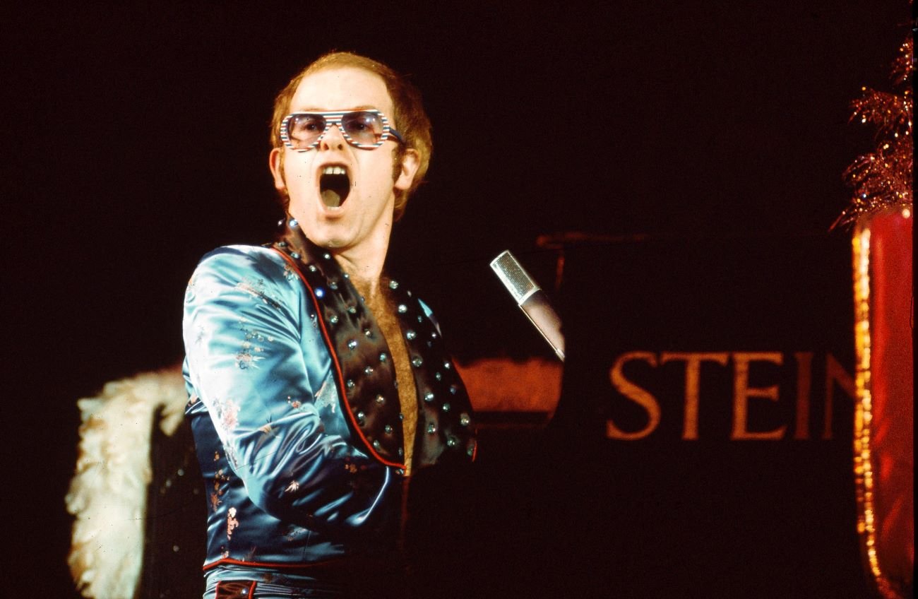 Elton John sits at a piano wearing a blue jacket and sunglasses in the 70s.