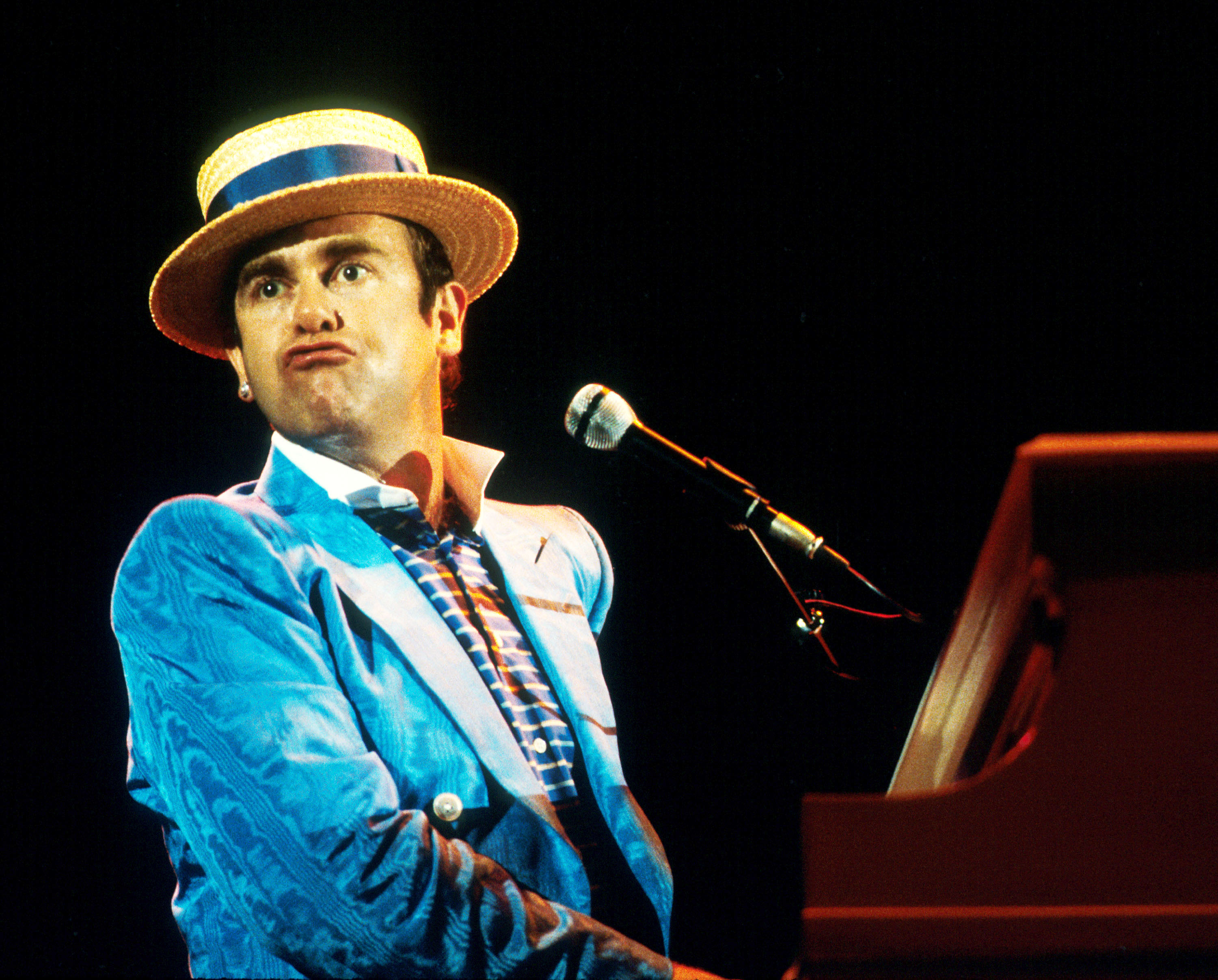 Elton John wears a blue coat and hat and sits at a piano.
