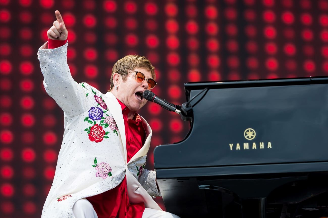 Elton John wears a white floral suit and plays the piano.