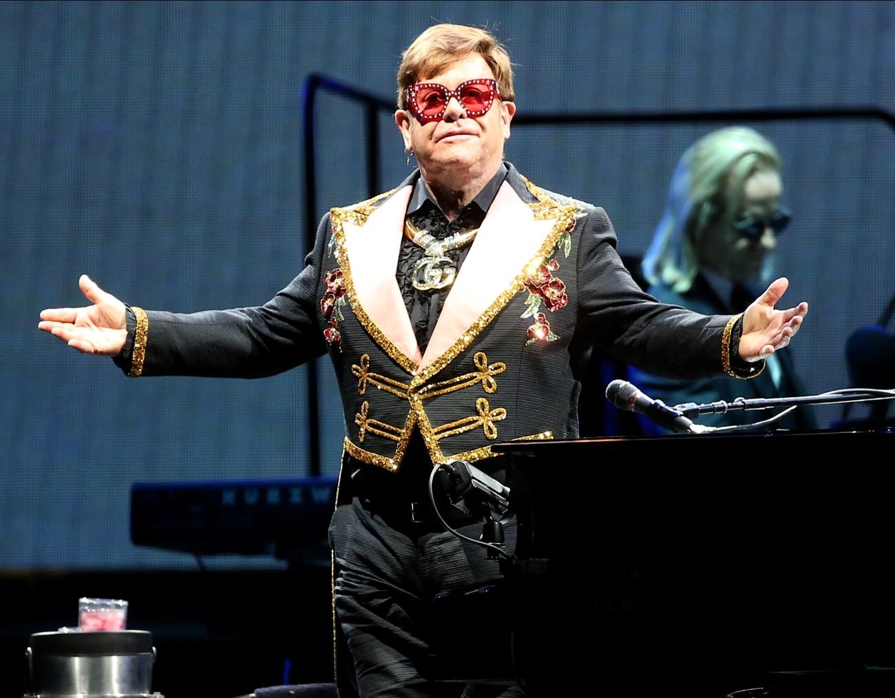 Elton John wears a black embroidered suit and red sunglasses. He stands in front of a piano.