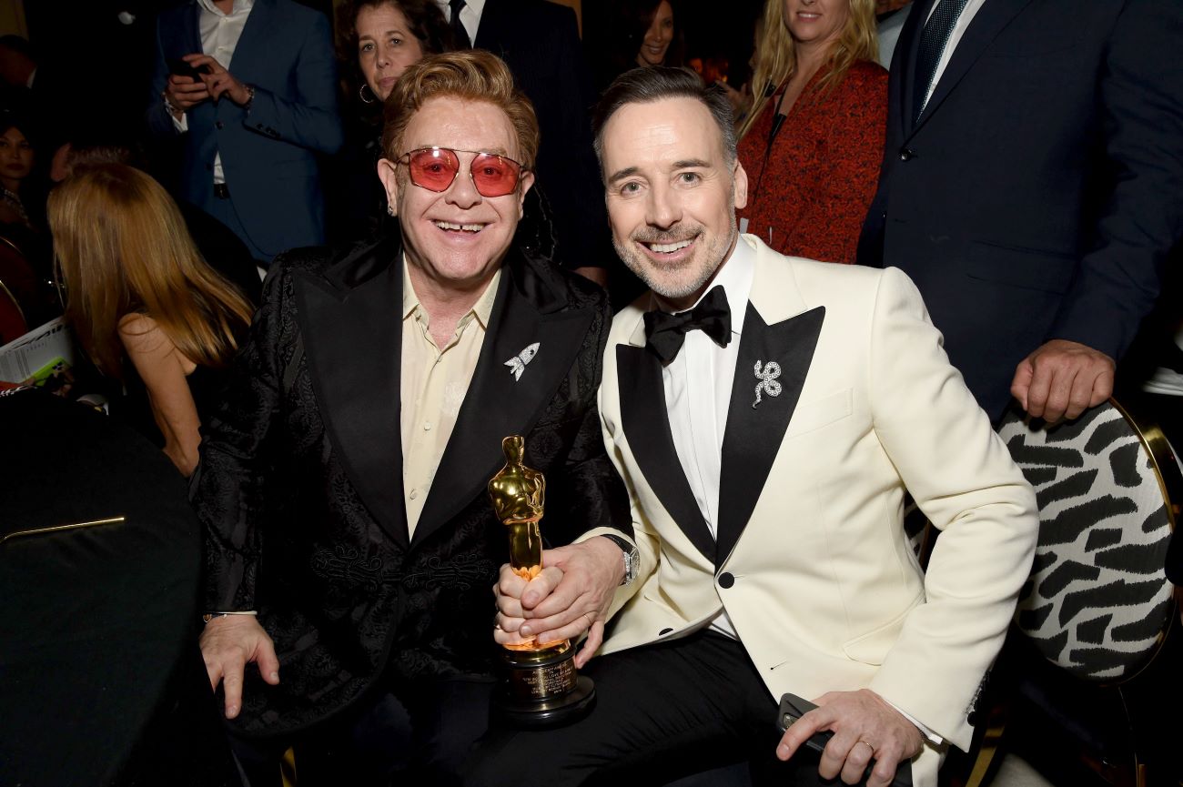 Elton John wears sunglasses and a black suit. David Furnish wears a white suit. They both hold an Oscar statue. 