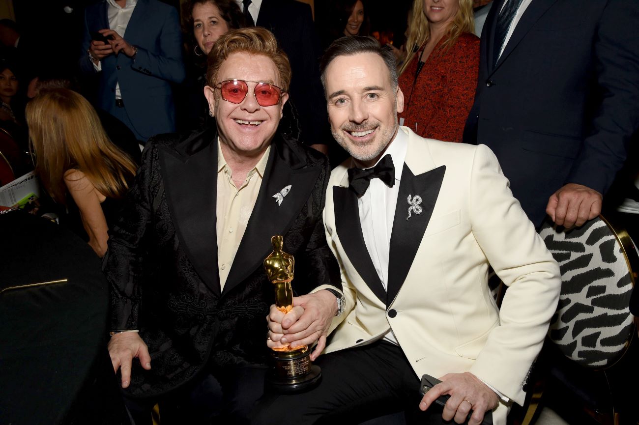 Elton John wears sunglasses and a black suit. David Furnish wears a white suit. They both hold an Oscar statue.