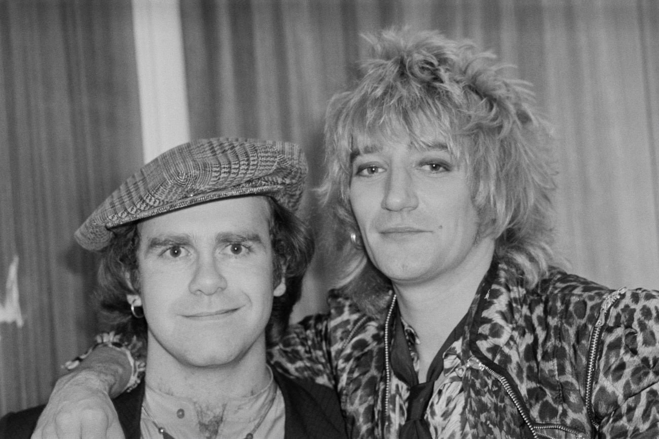 A black and white photo of Elton John wearing a hat and Rod Stewart wearing a cheetah print coat.