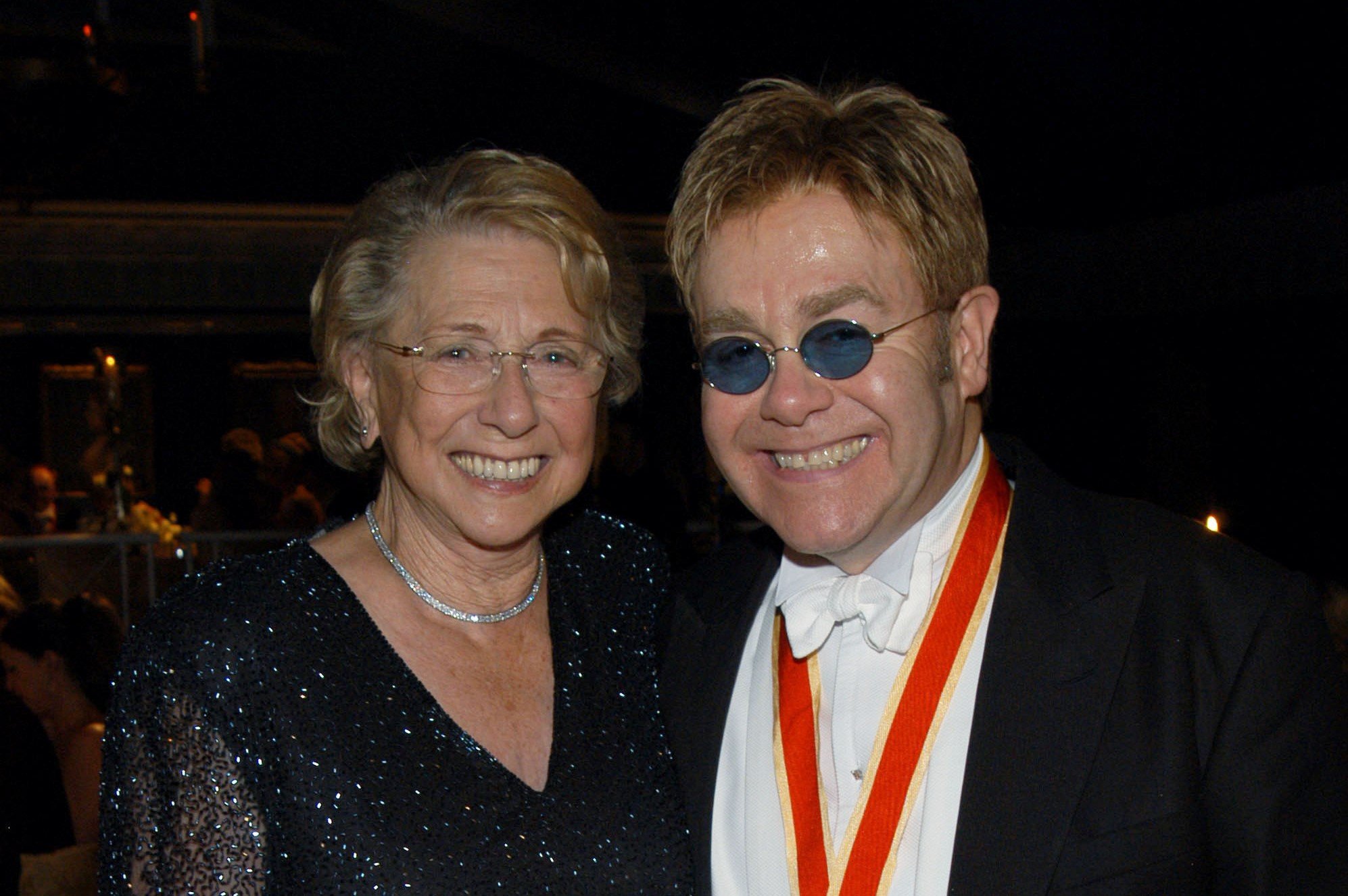 Elton John and his mom pose smiling together. Elton John's mom wears a black sparkling dress and he wears a tuxedo and sunglasses.