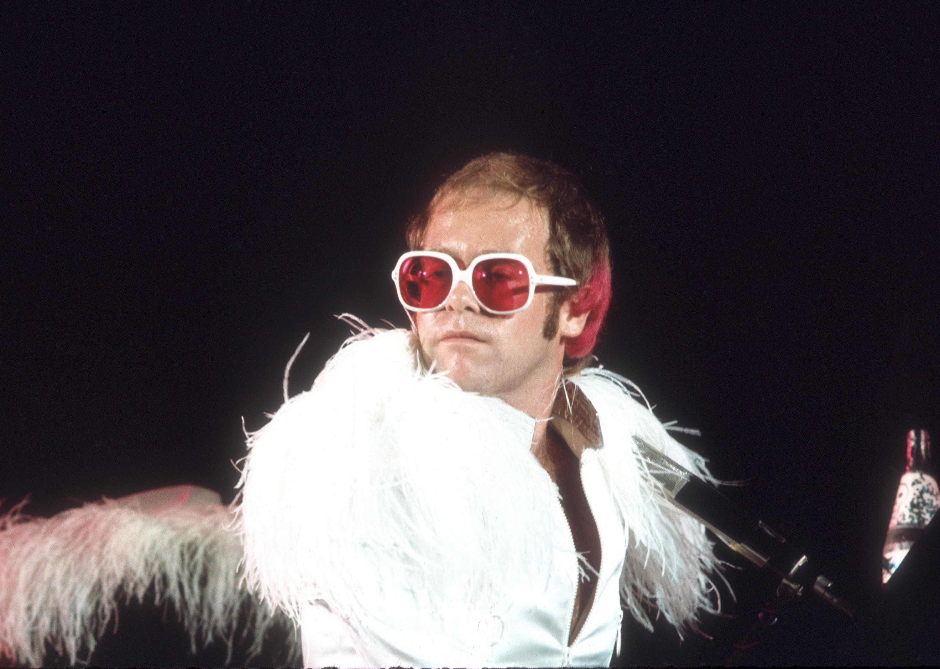 Elton John wears white and red sunglasses and a white feather coat.