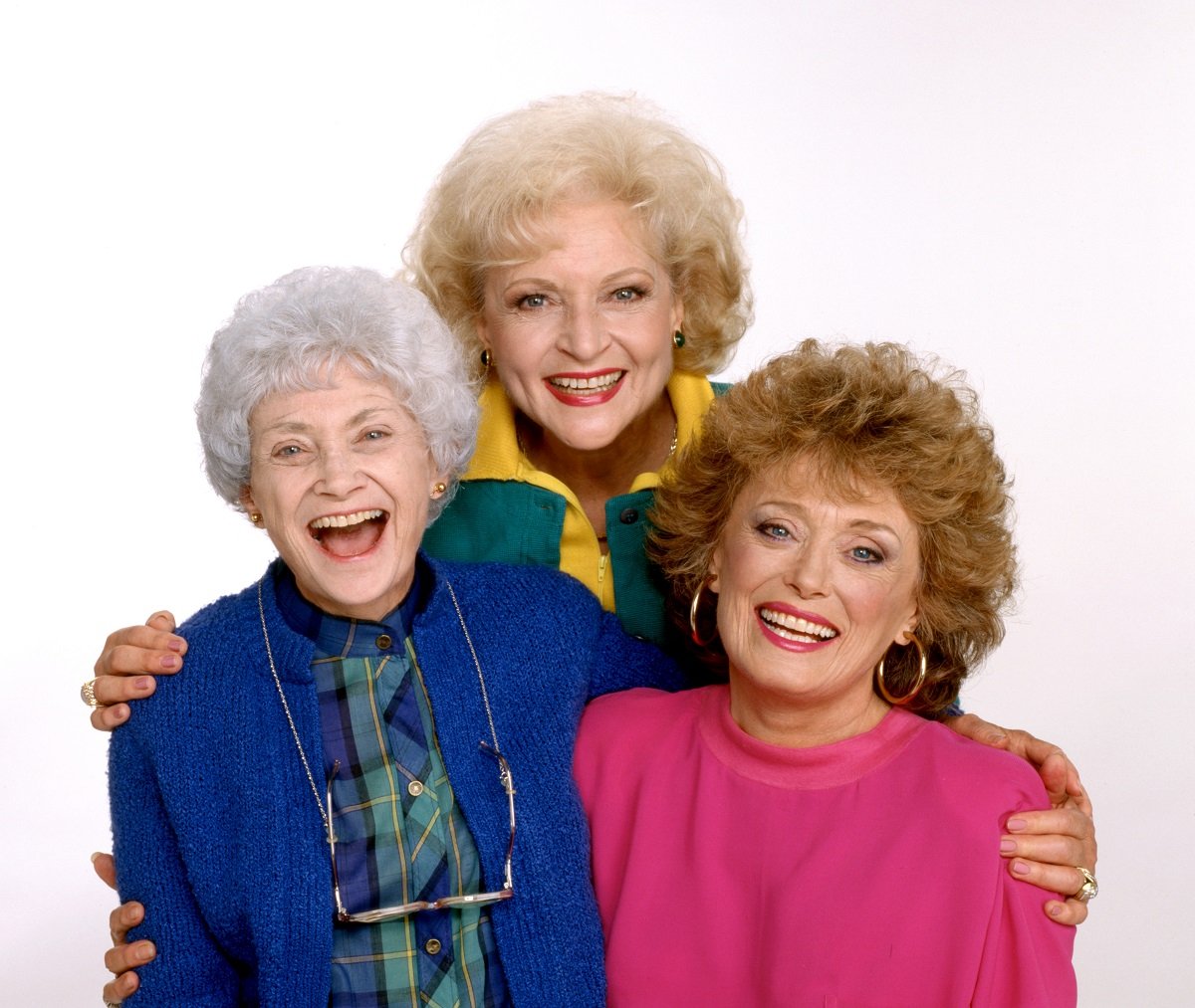 'The Golden Girls' actor Estelle Getty in a blue dress, Betty White in green and yellow outfit, and Rue McClanahan in a pink sweater; pose in front of a white backdrop.
