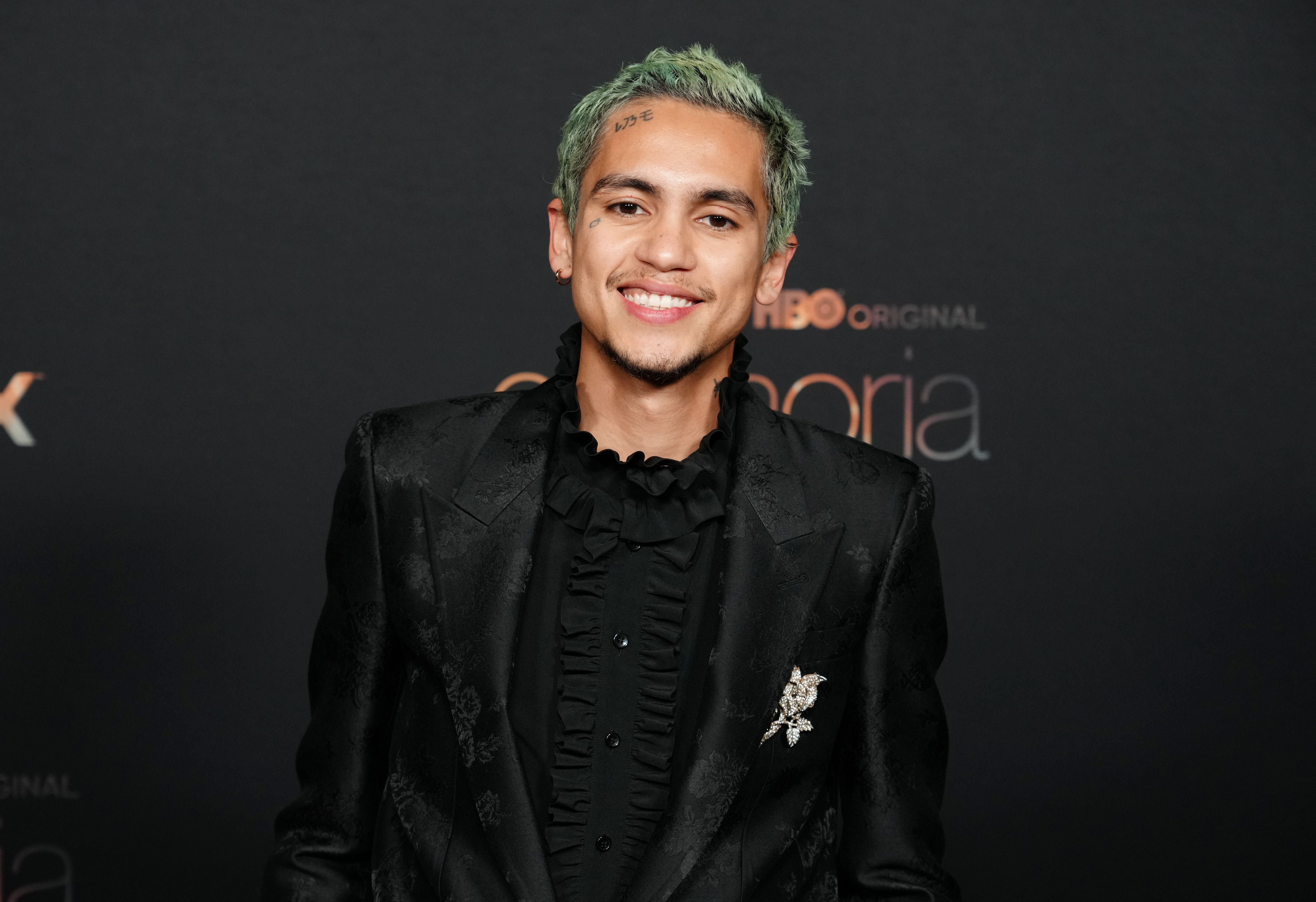 Elliot actor Dominic Fike attends HBO's Euphoria Season 2 Photo Call. Fike has green hair and wears a black suit jacket.