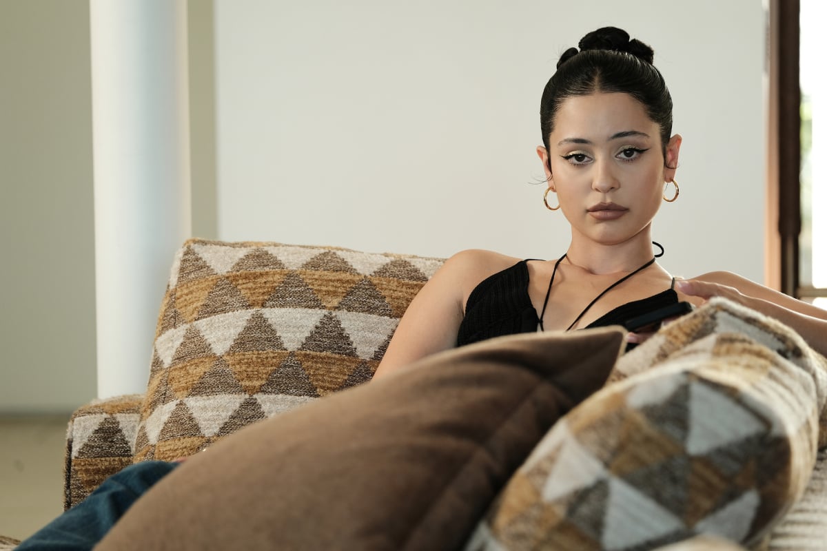 Alexa Demie as Maddy in Euphoria Season 2. Maddy sits on the couch looking apathetic. She is wearing gold hoop earrings and has her hair in a bun.