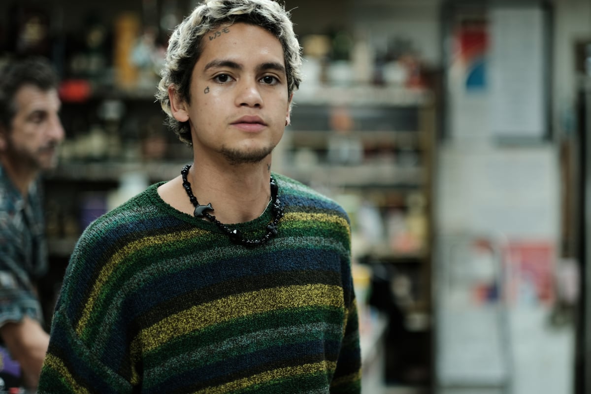 Dominic Fike as Elliot in Euphoria Season 2. Elliot wears a necklace and striped sweater and stands in a convenience store.