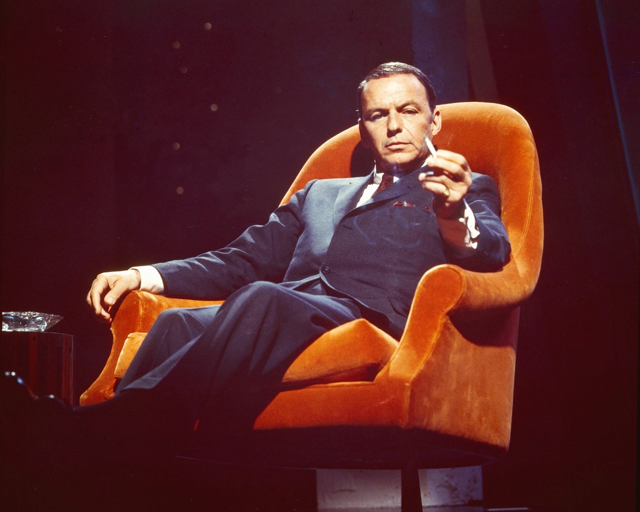 Frank Sinatra wears a suit and sits in an orange chair with a cigarette.