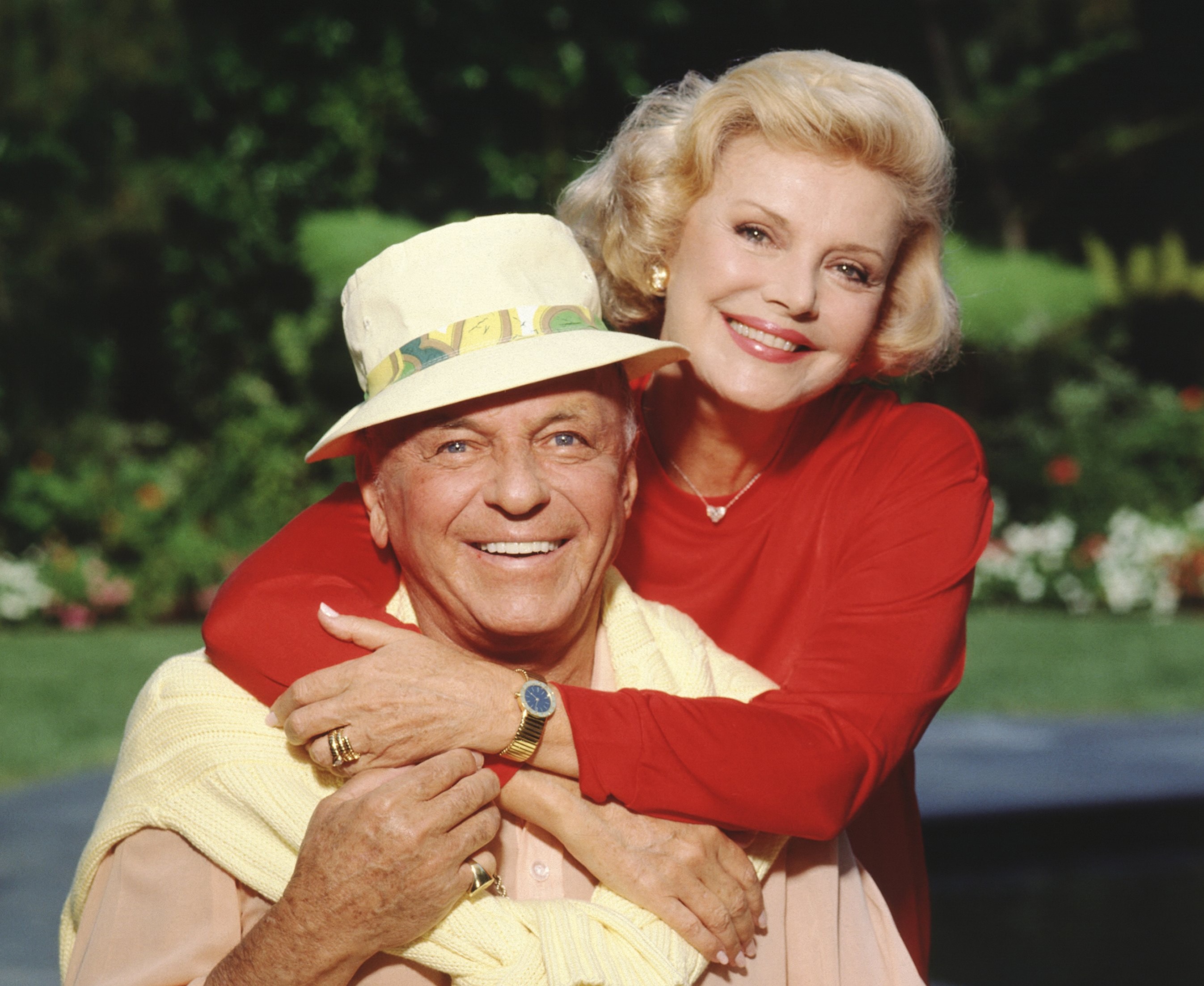 Frank Sinatra wears a yellow hat and sweater. Frank Sinatra's spouse Barbara Sinatra wears a red sweater and wraps her arms around him.