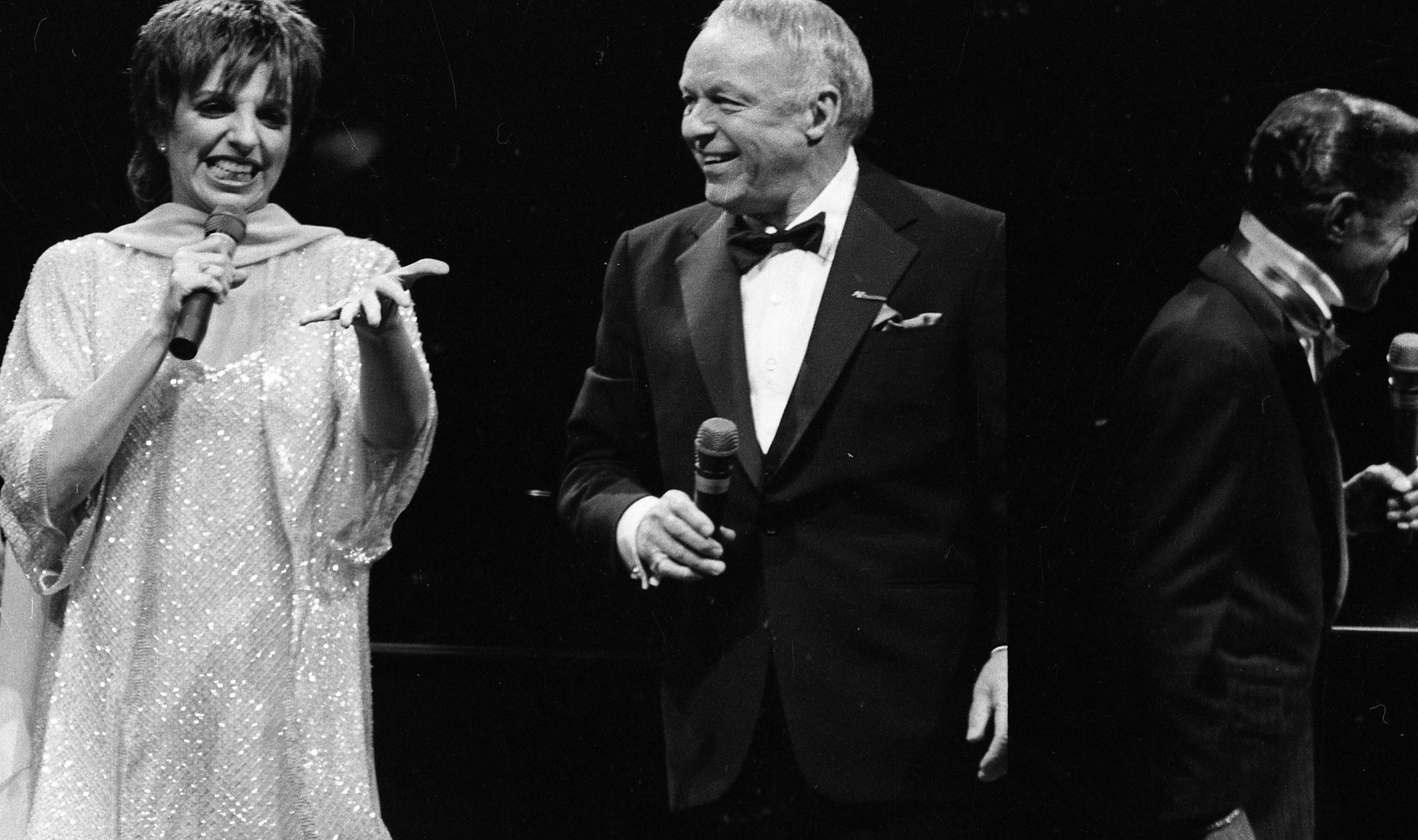 A black and white photo of Liza Minnelli and Frank Sinatra holding microphones. Sammy Davis Jr. faces away from the camera.