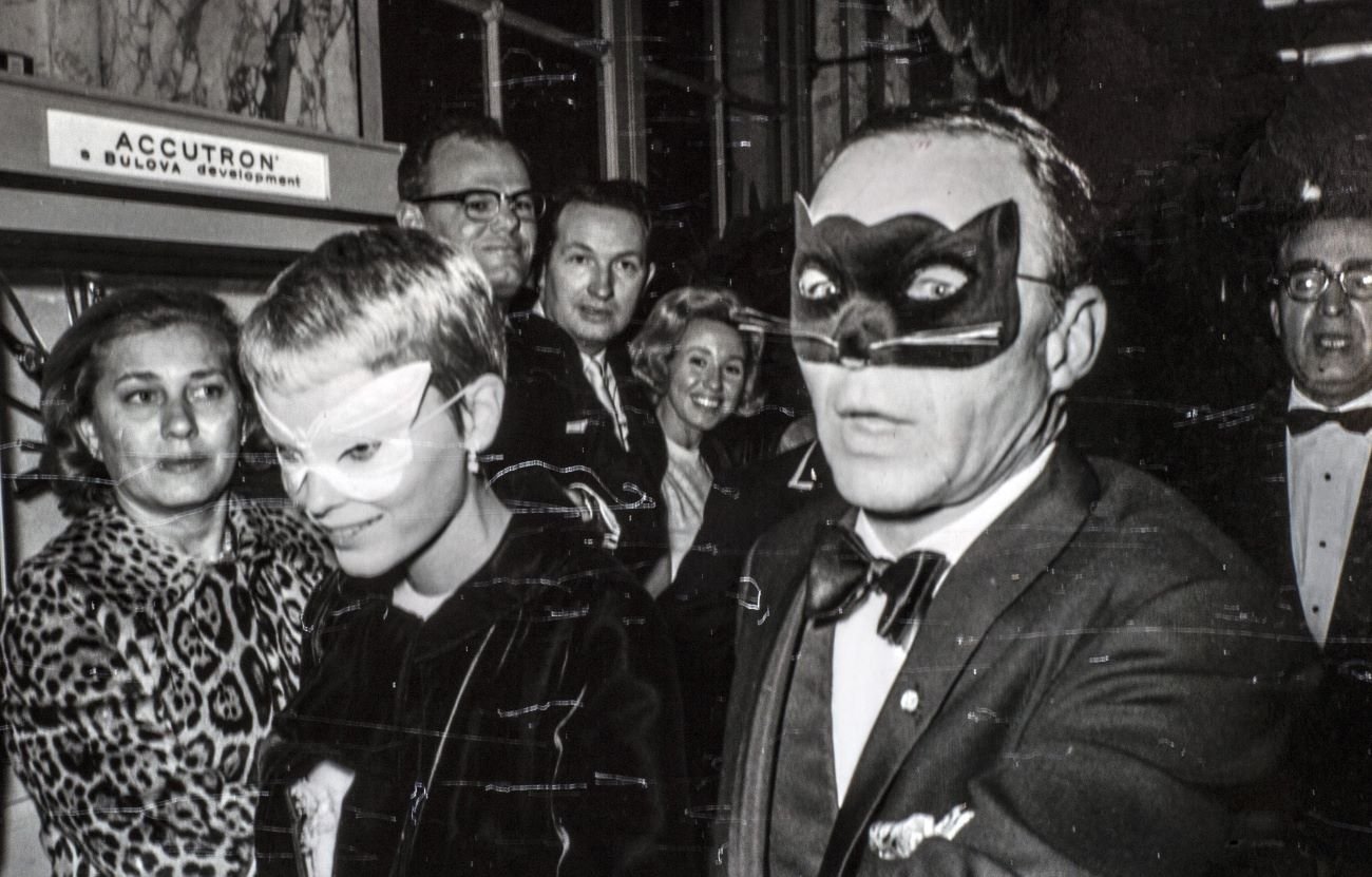 Frank Sinatra and Mia Farrow walk through a crowd. She wears a white butterfly mask and he wears a black cat mask.