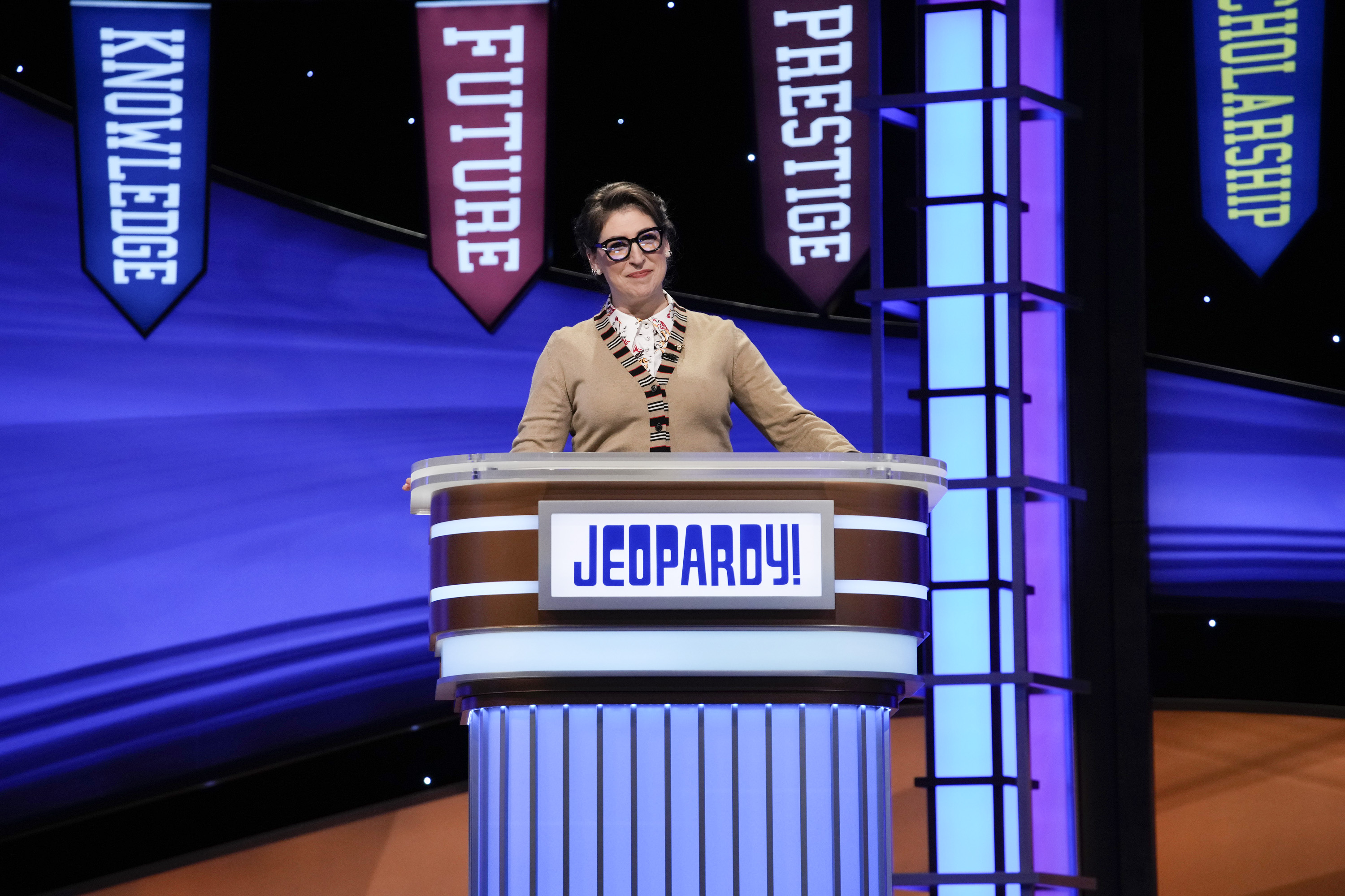 'Jeopardy!' host Mayim Bialik wears a brown sweater and glasses at the lectern for the show's National College Championship tournament.