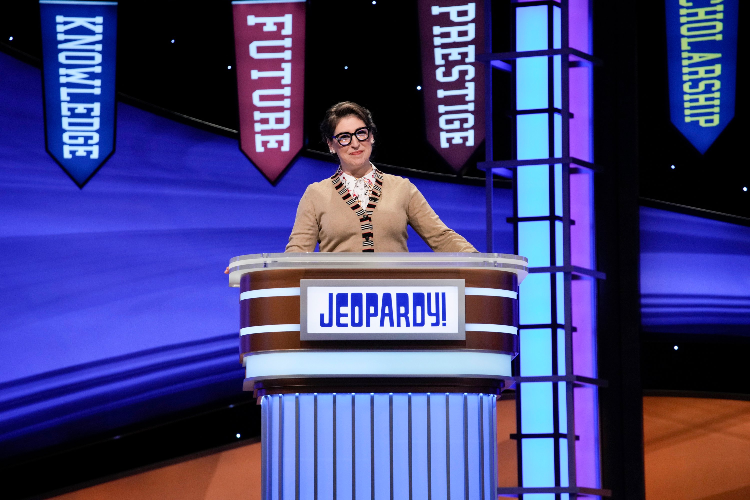 'Jeopardy!' host Mayim Bialik wears a brown sweater and glasses at the lectern for the show's National College Championship tournament.